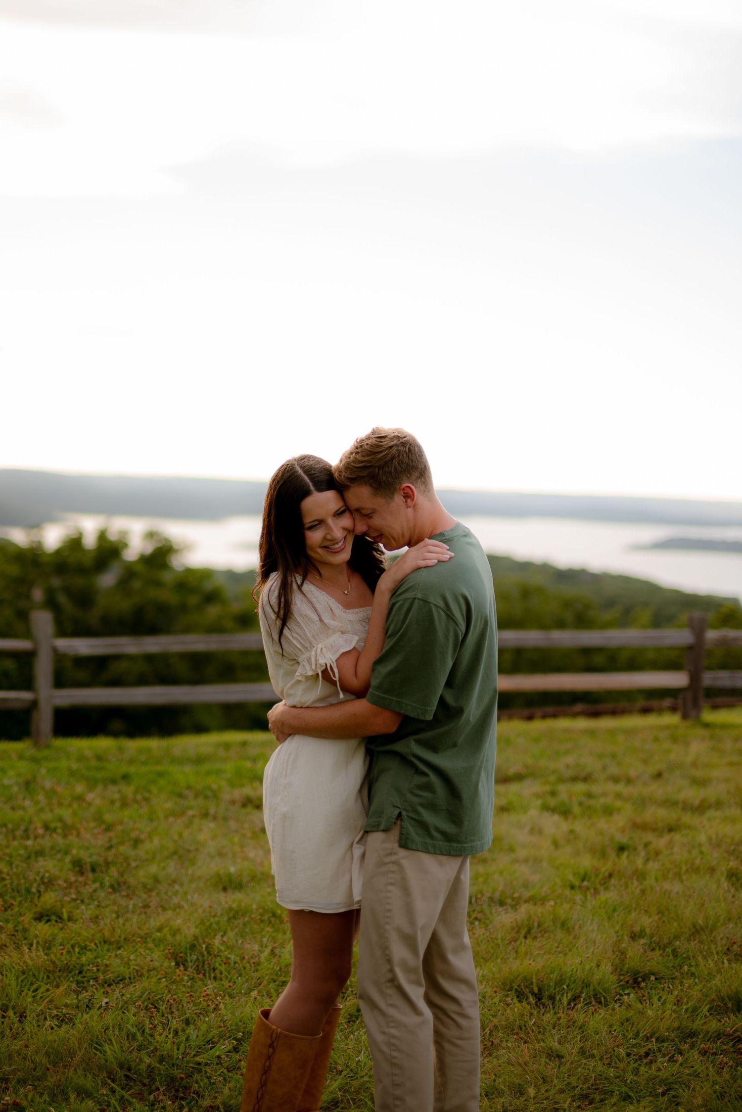A couple embraces tenderly on a grassy hilltop with a scenic lake and lush greenery in the background.