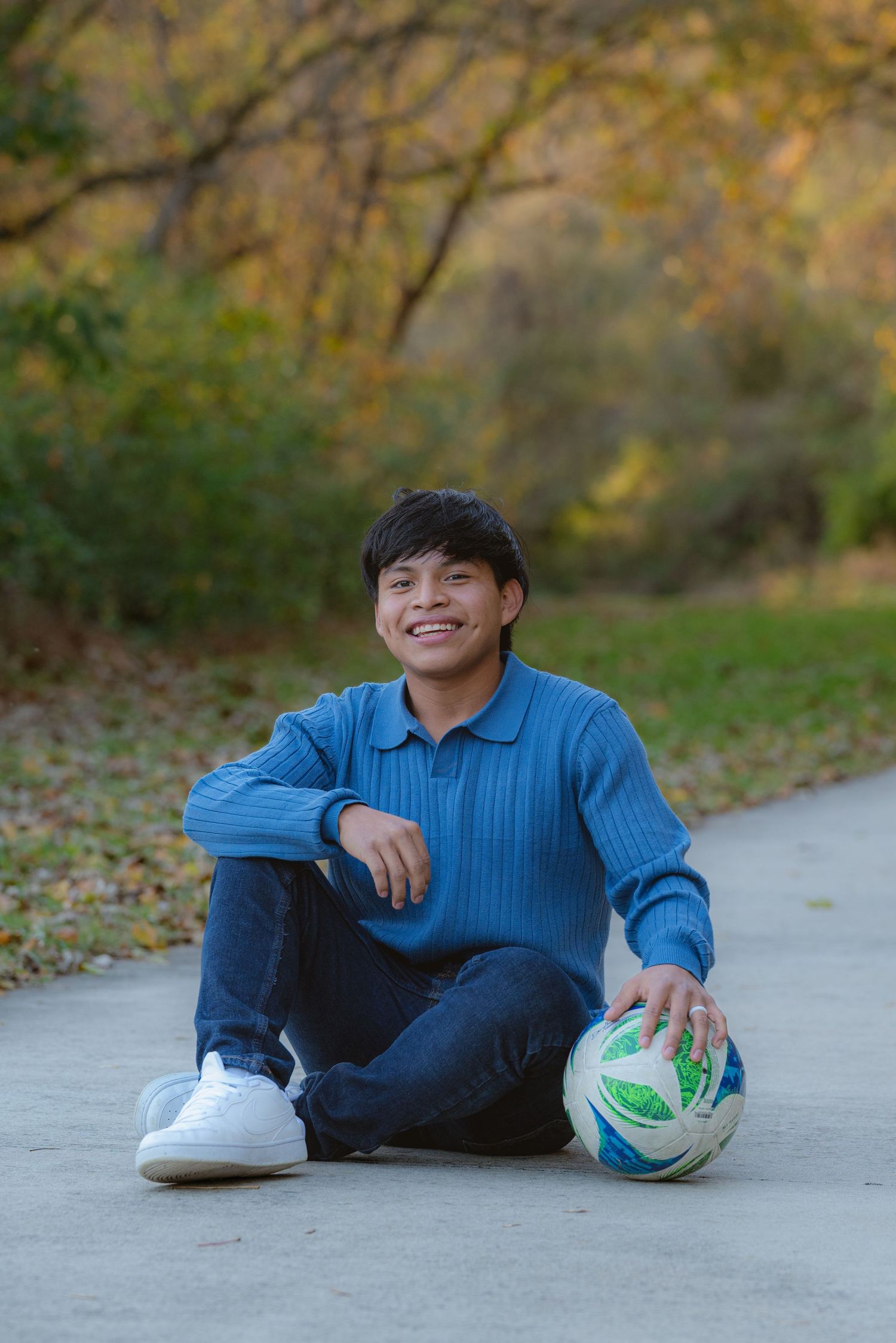 Smiling teen boy in blue sweater sitting on path with soccer ball in fall park.