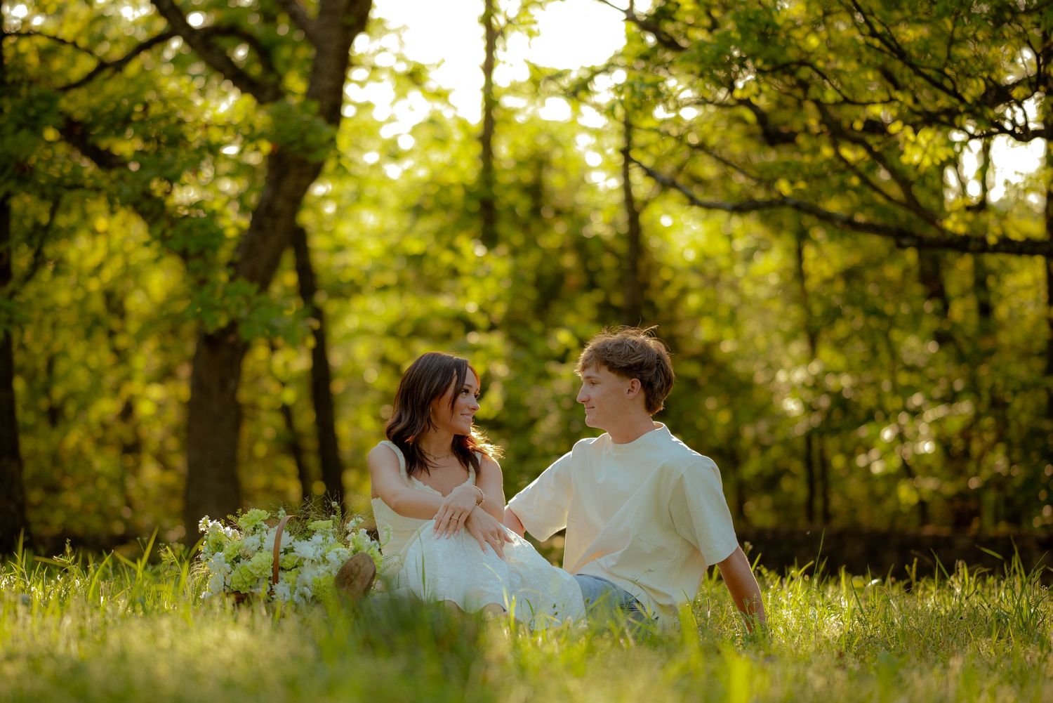 Couple sitting together in a sunlit meadow surrounded by lush green trees, sharing a romantic moment outdoors.