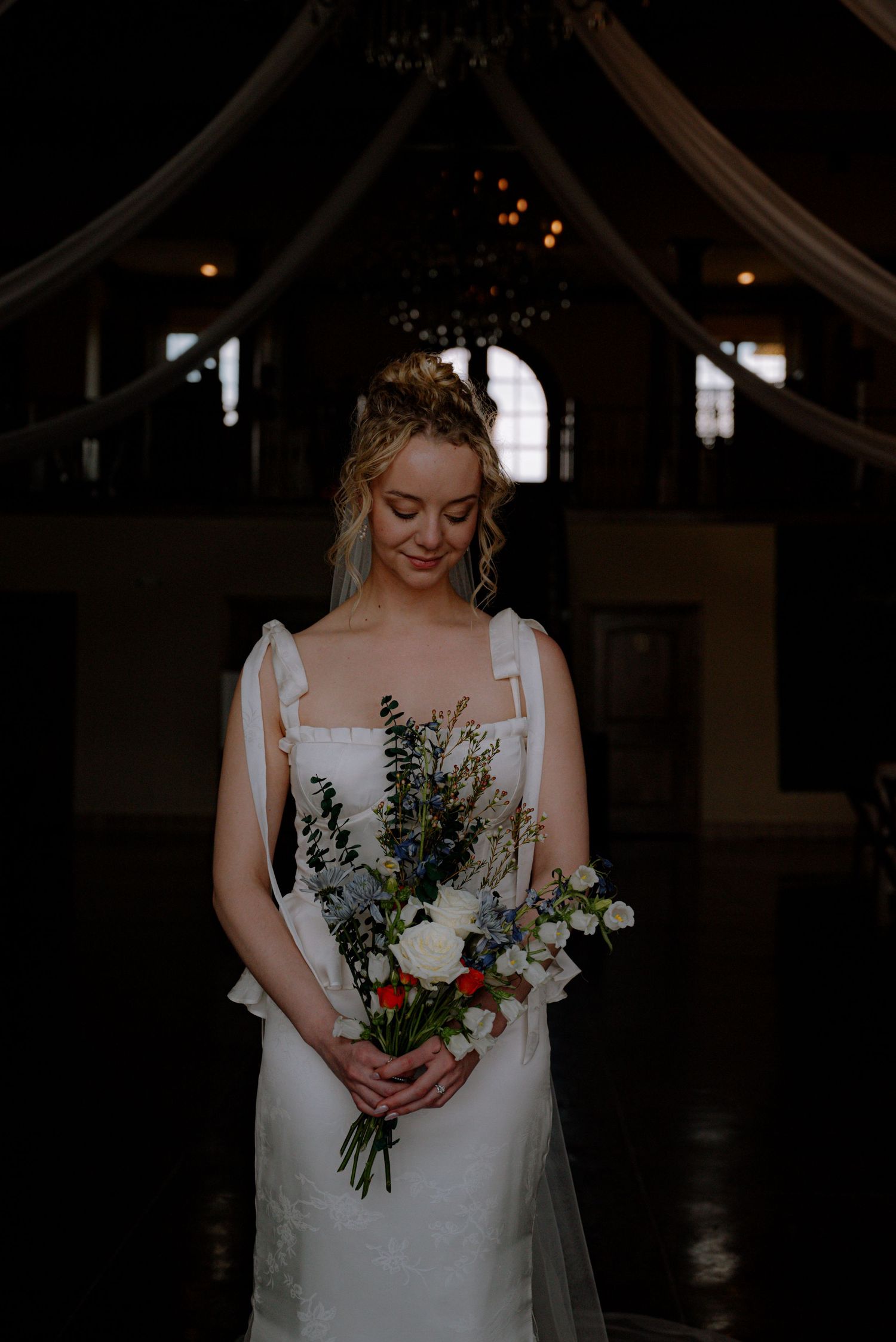 Blonde bride in lace gown holds a wildflower bouquet with blue and white blooms in a dark venue.