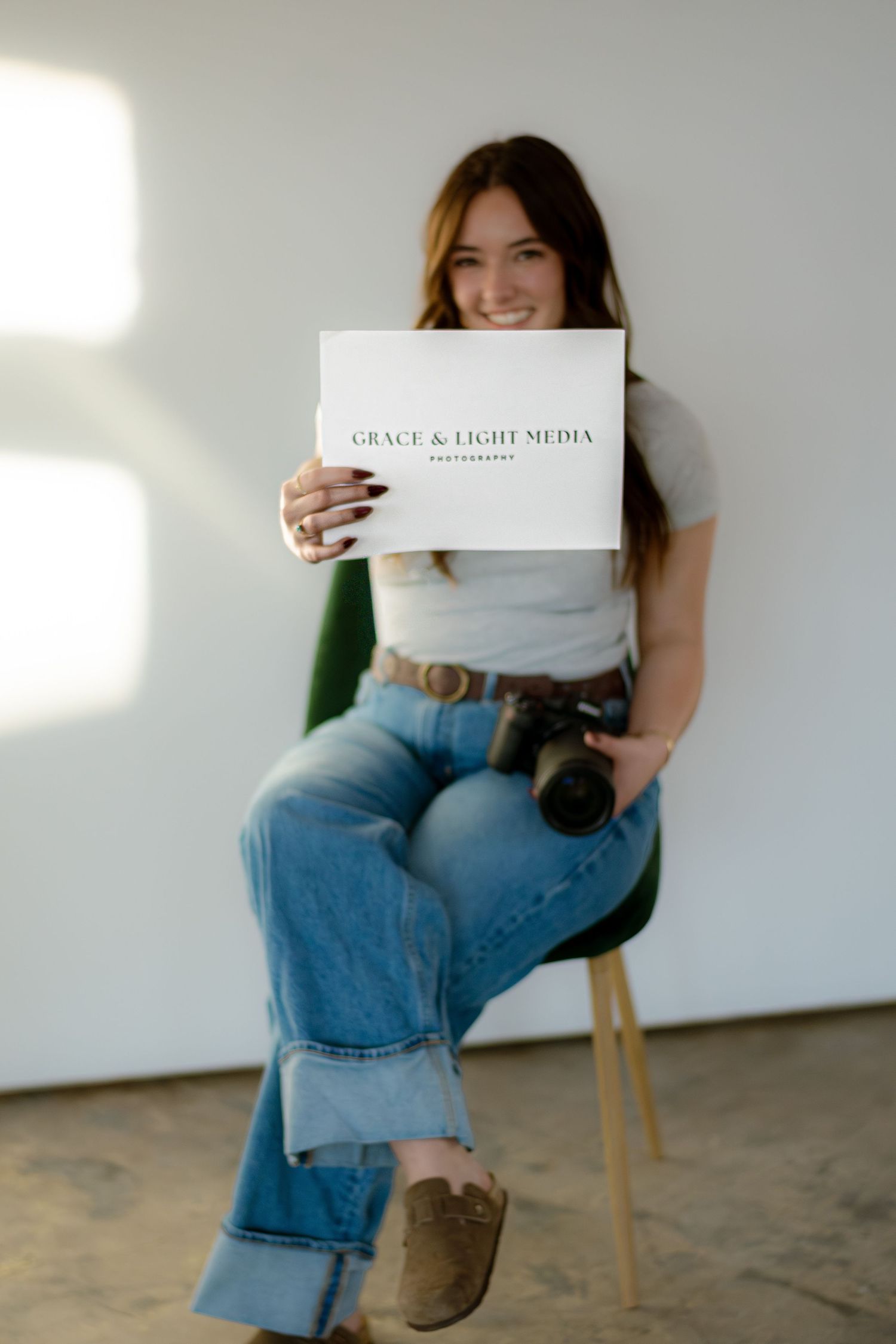 Smiling woman sitting in green chair holding Grace & Light Media Photography sign with camera in hand.