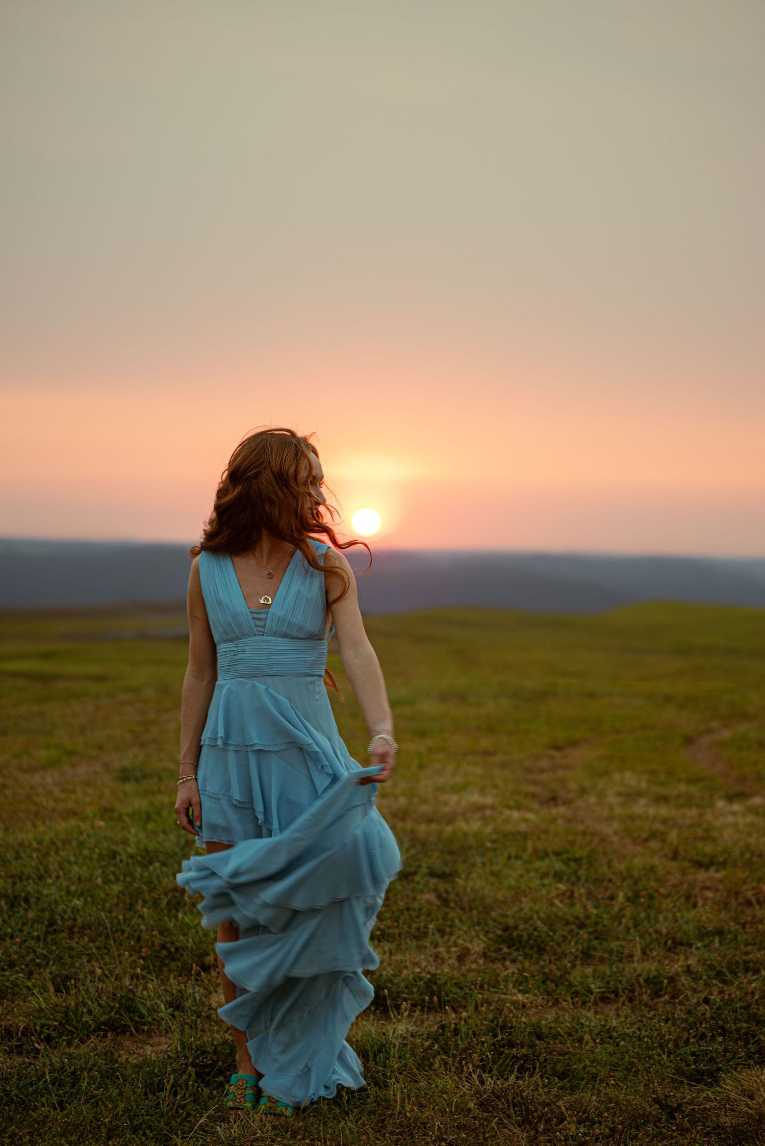 Woman in flowing teal tiered dress walking through open field toward vibrant pink sunset.