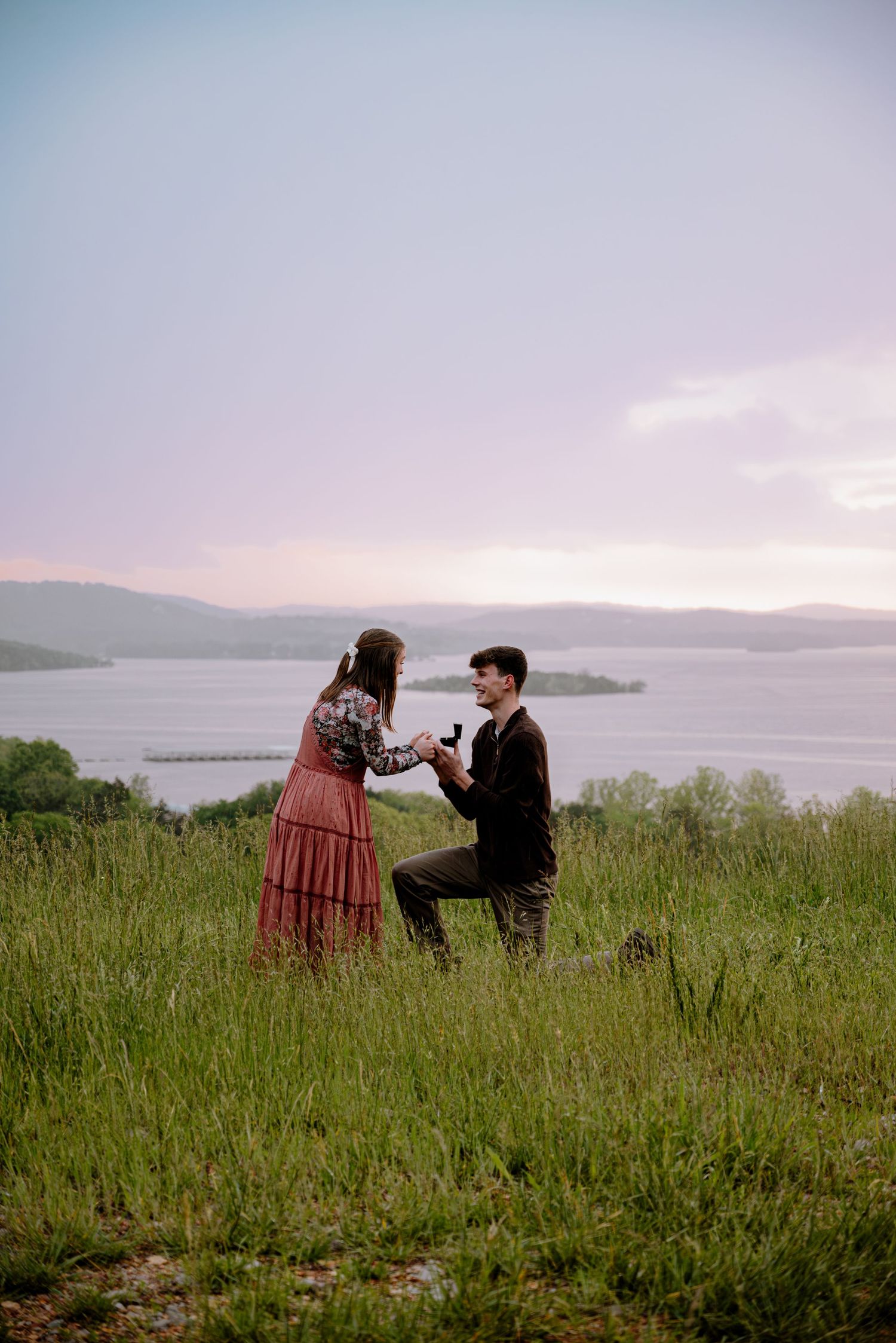 Romantic marriage proposal at sunset with man on one knee holding ring box in a scenic lakeside field.
