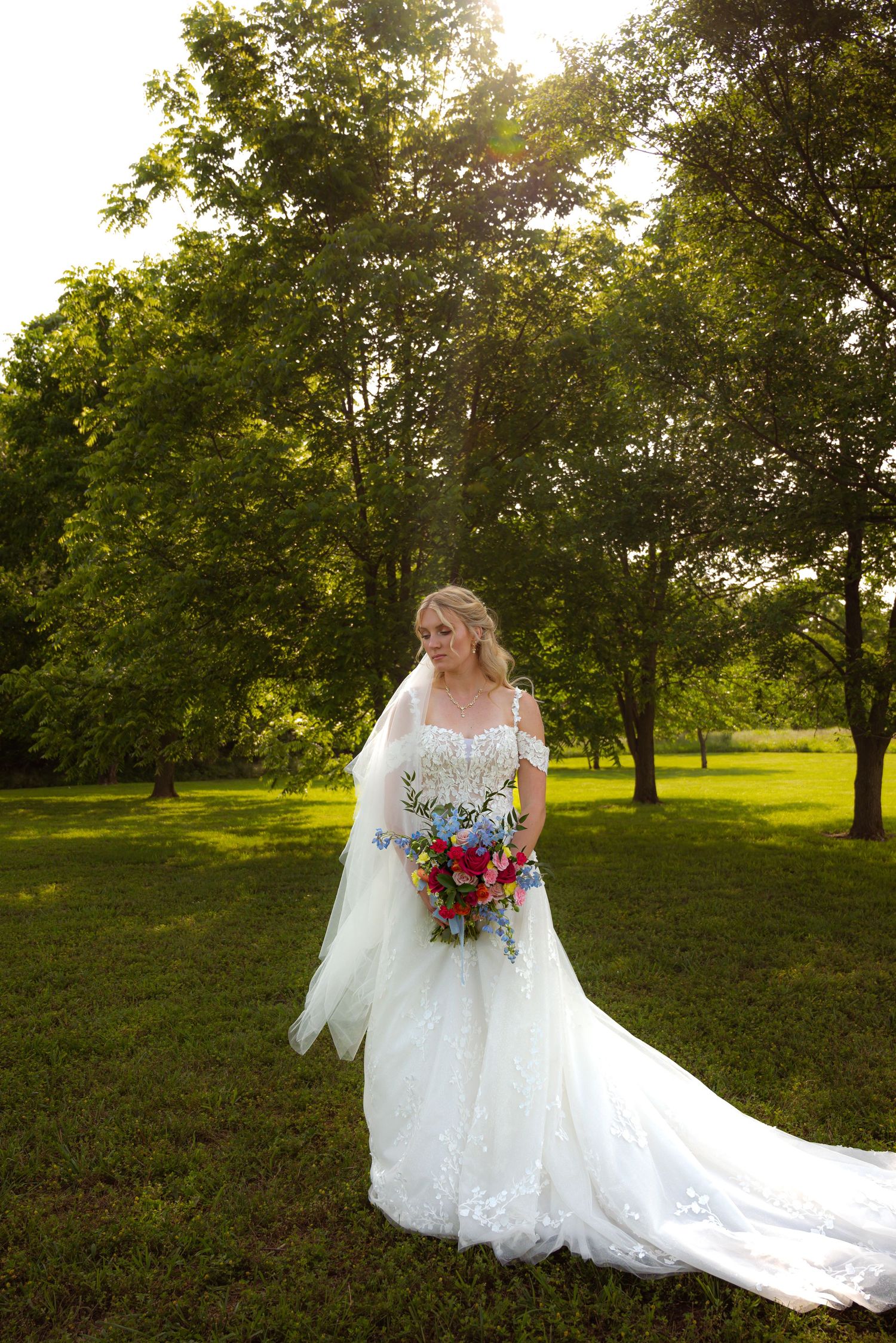Bride in lace wedding gown holding colorful bouquet standing in sunlit green garden.