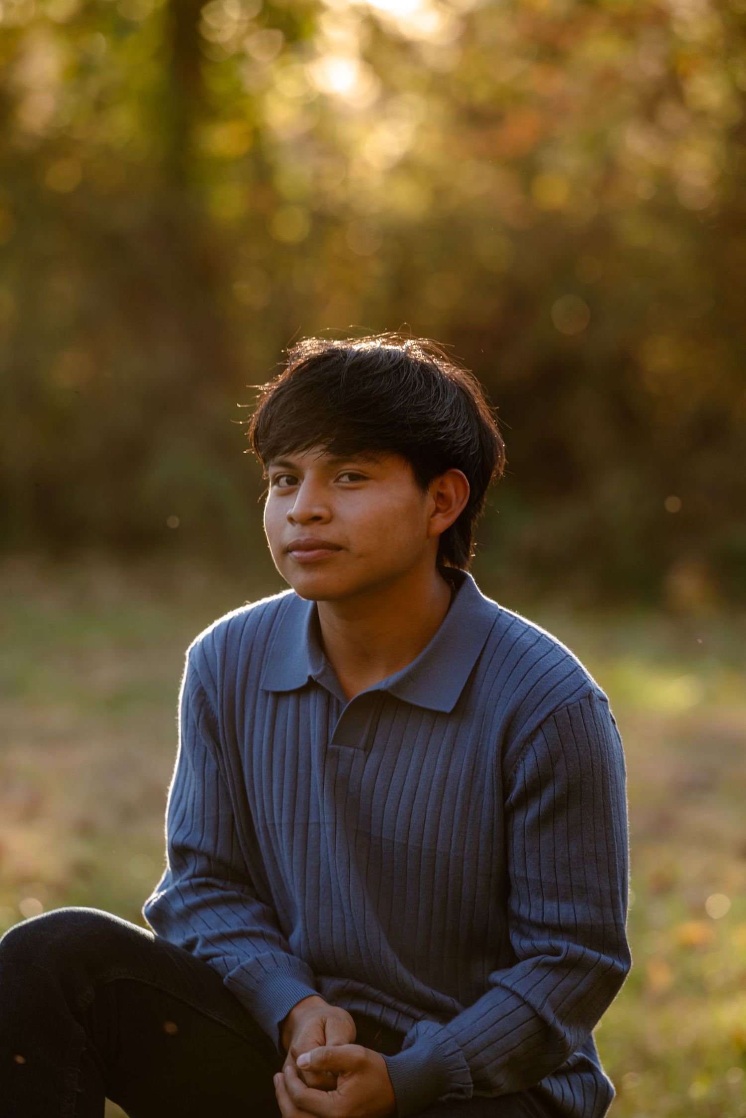 Young man in blue ribbed polo sitting outdoors in golden autumn light looking away.