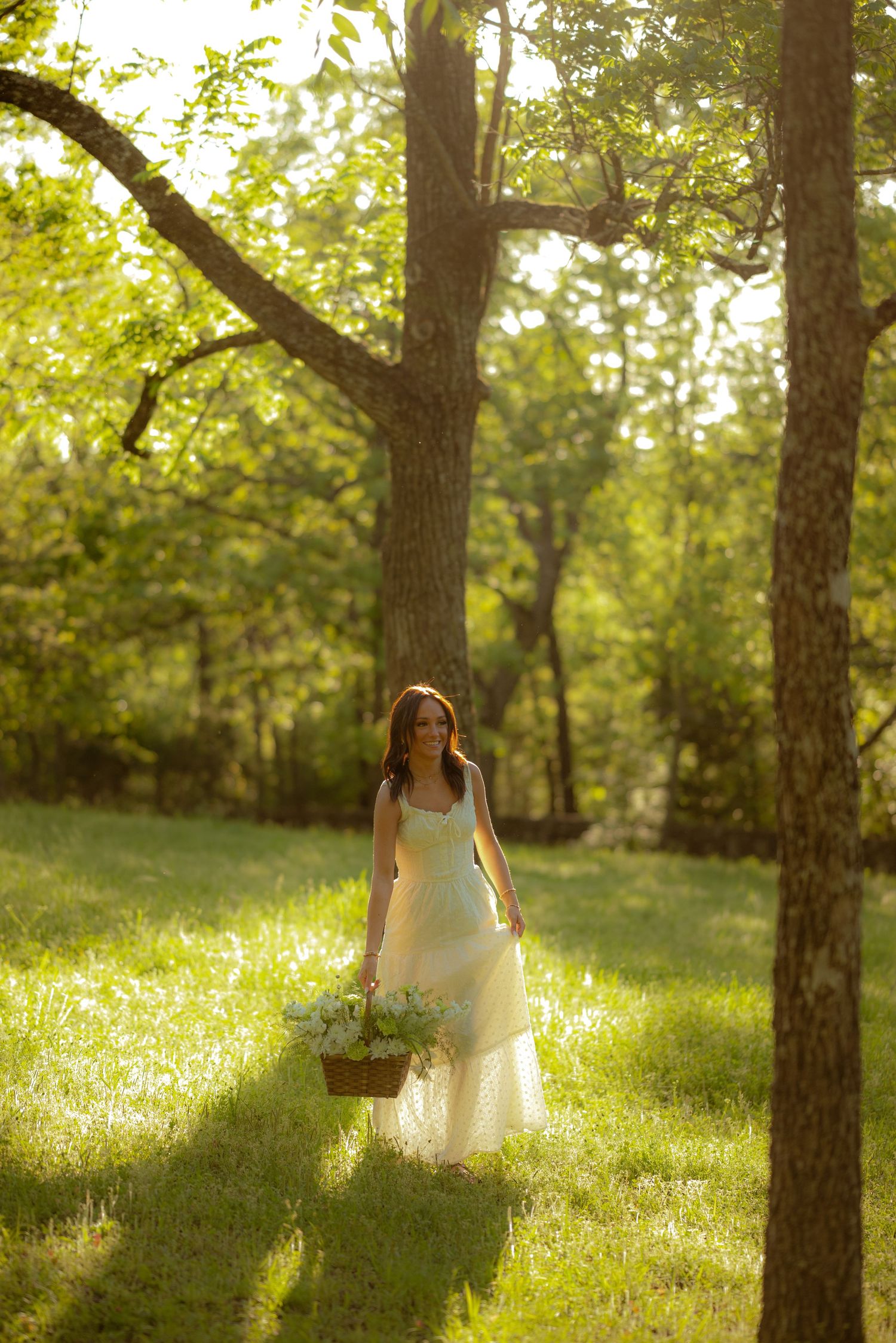 Woman in white dress carrying flower basket walks through sunlit green forest clearing.