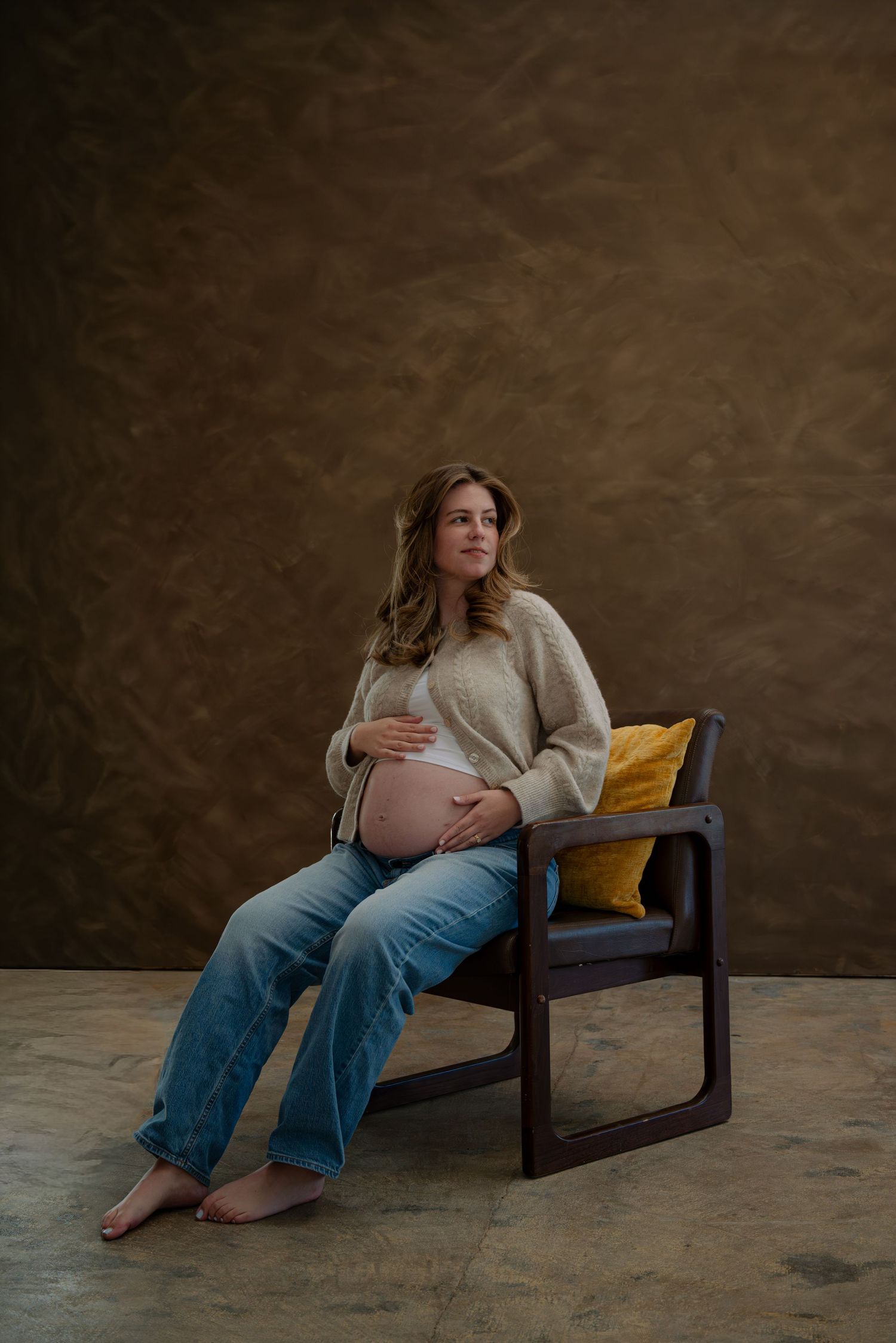 Pregnant woman in beige sweater and jeans sitting in dark chair with yellow pillow in studio.
