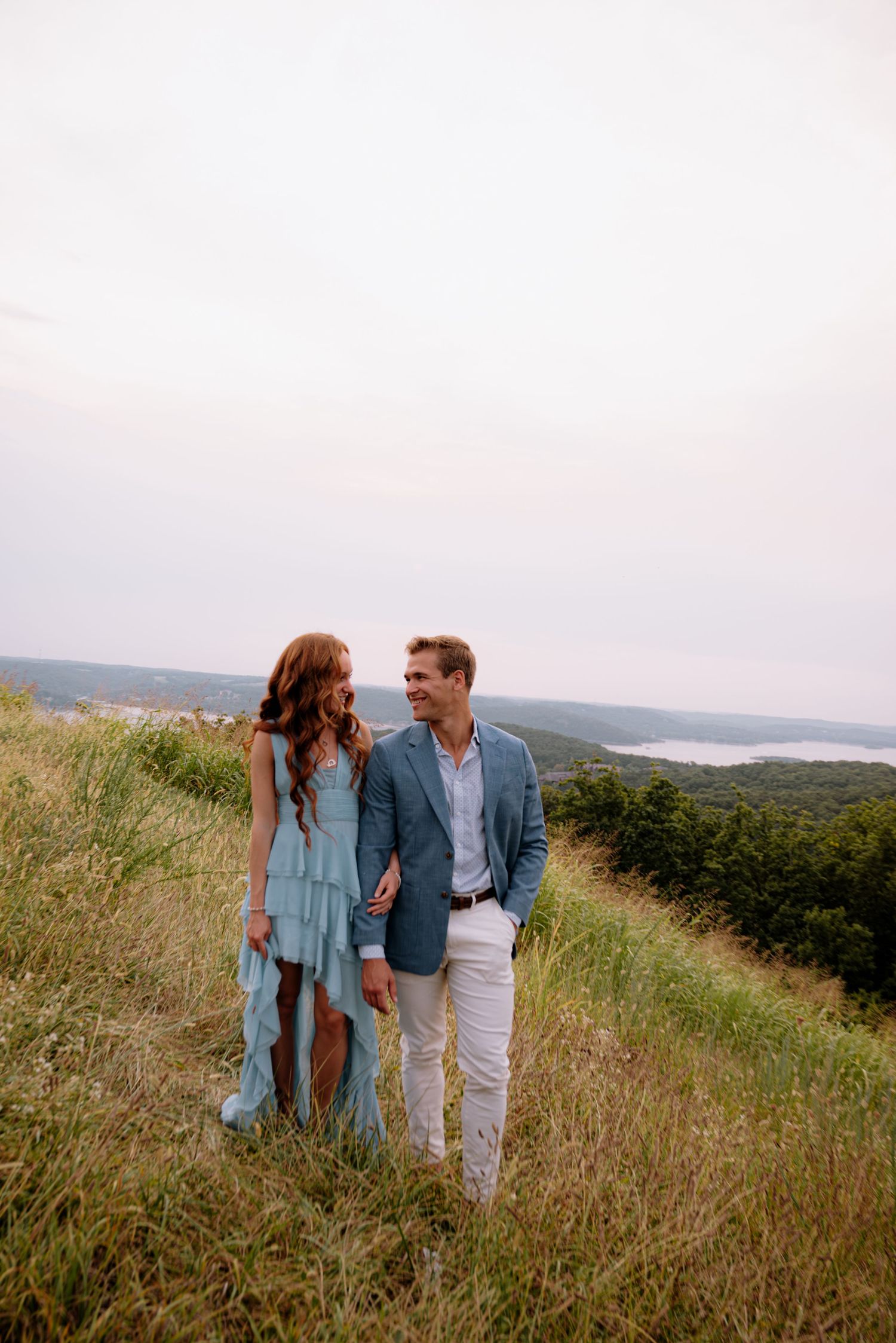 Couple in coordinating blue outfits walking hand-in-hand on a grassy hilltop with scenic landscape views.
