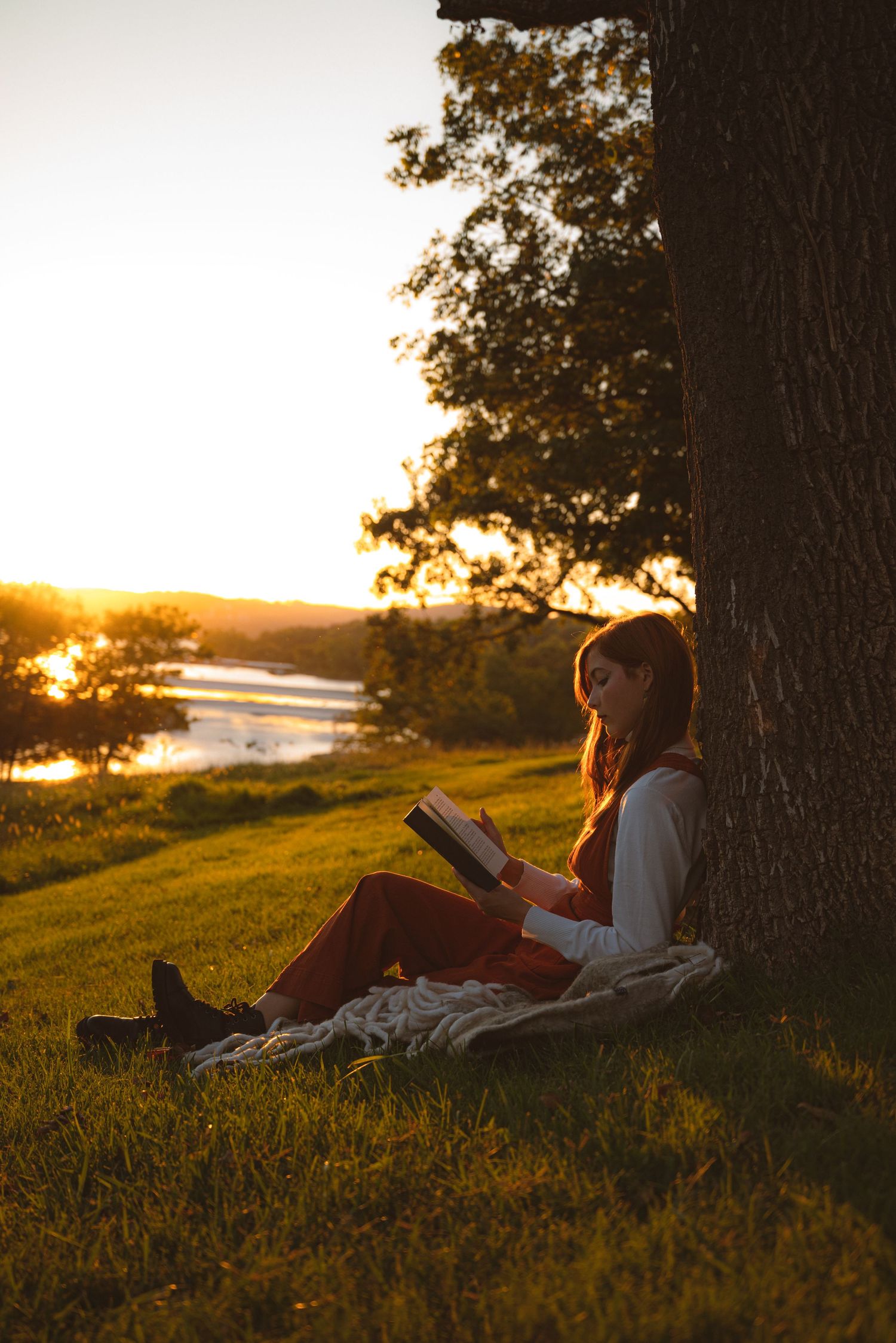 Young woman in rust outfit reads a book leaning against a tree at a scenic golden sunset.