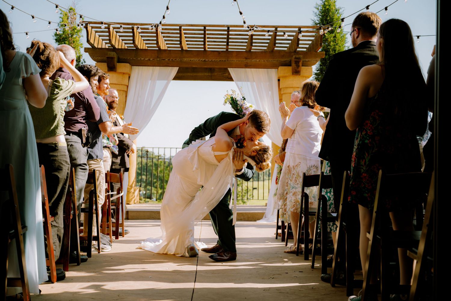 Bride and groom sharing a kiss at their outdoor ceremony, surrounded by cheering friends and family