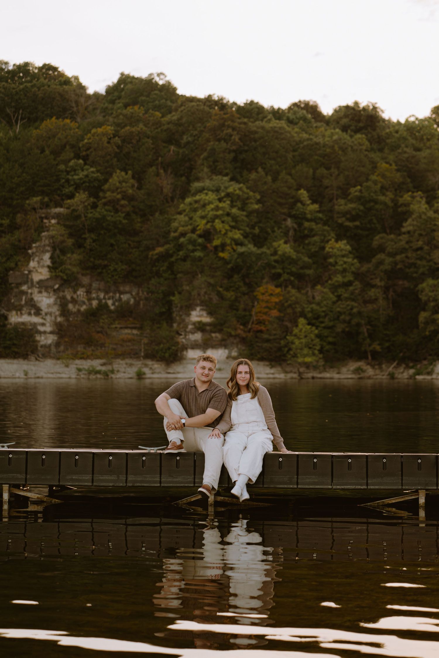 Couple sitting together on a wooden dock over a calm lake surrounded by autumn foliage.