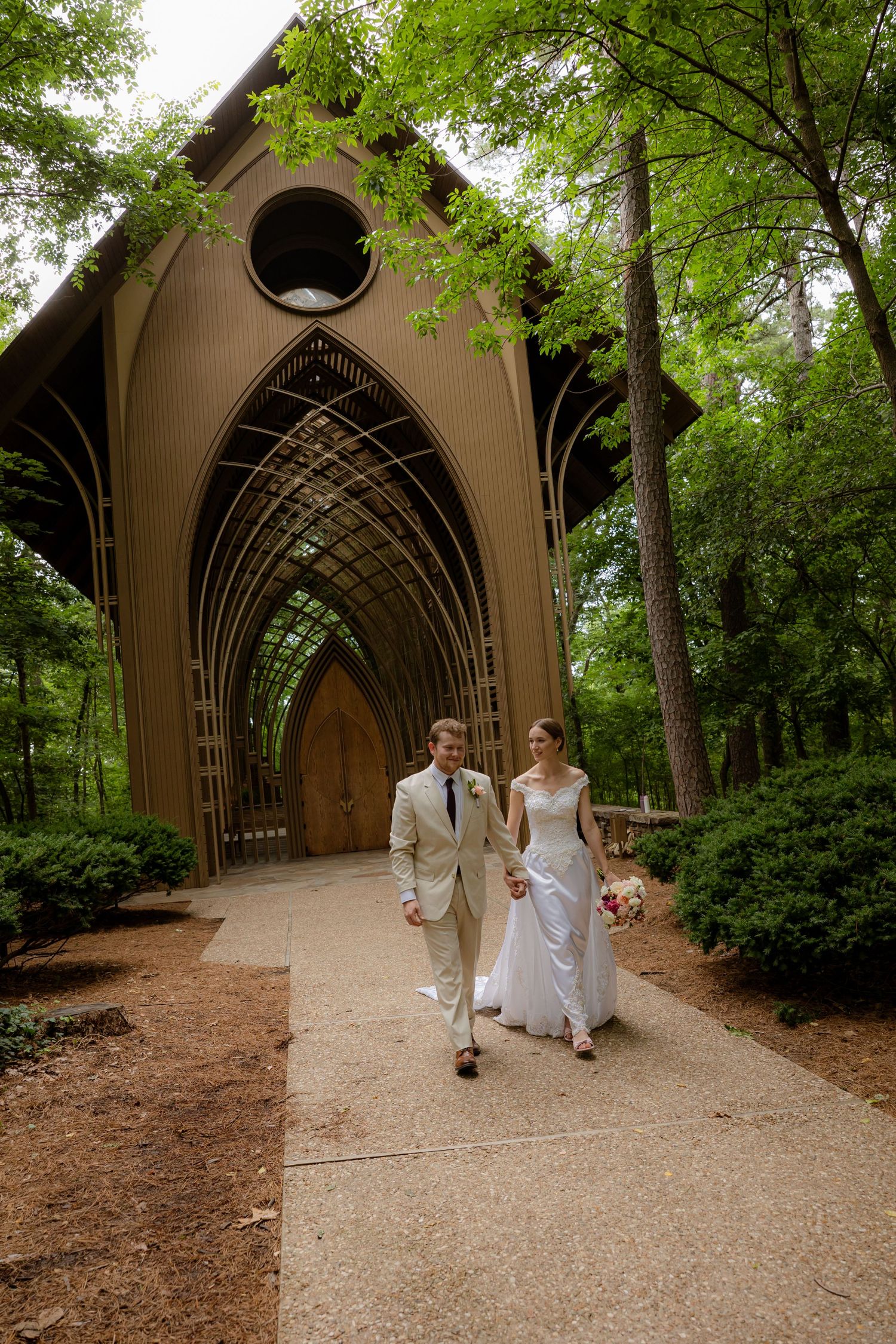 Bride and groom walk hand-in-hand outside the Mildred B. Memorial Chapel surrounded by lush green trees on their wedding day.