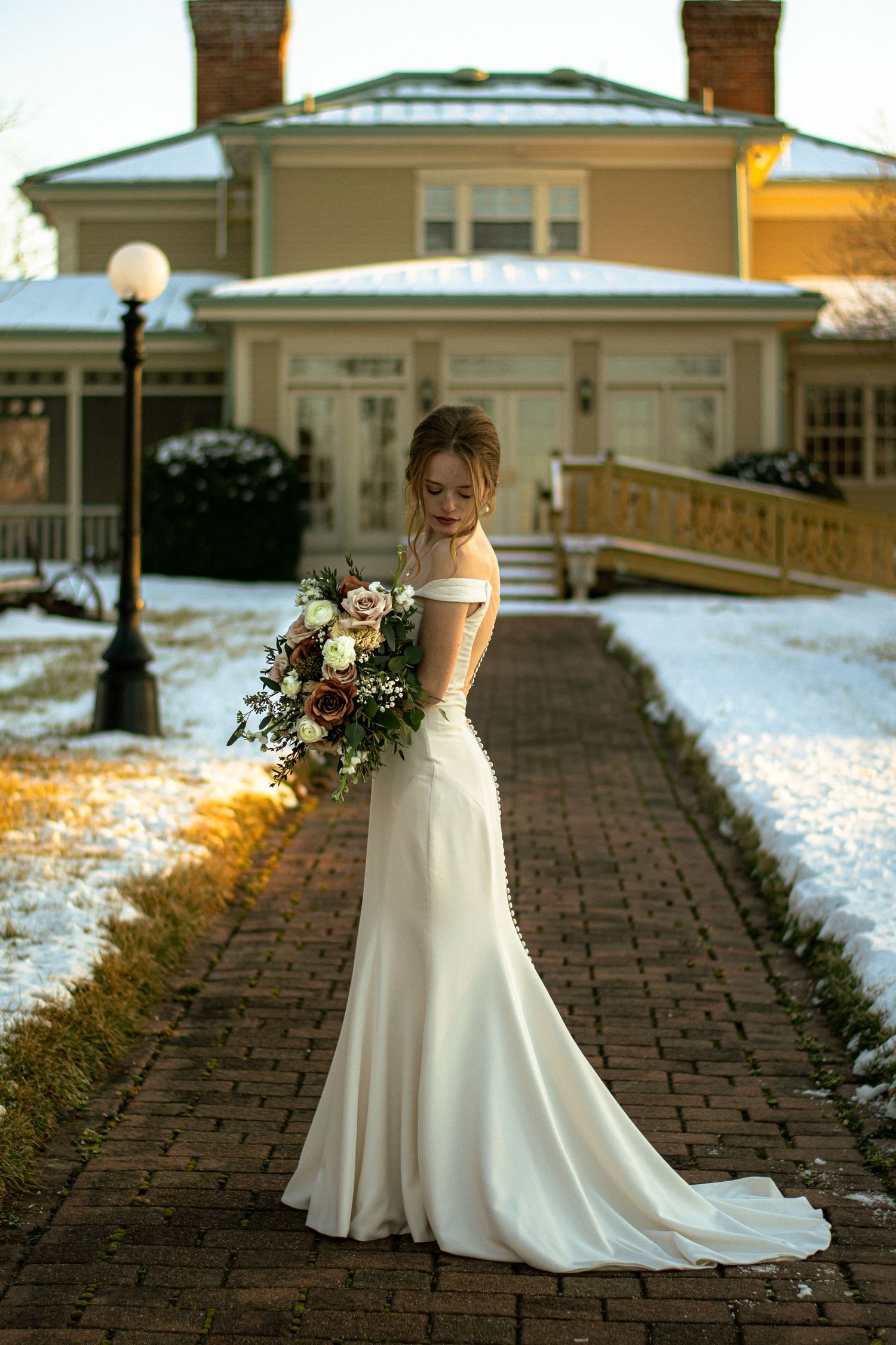 Elegant bride in fitted off-shoulder gown holds bouquet on snowy brick path at golden hour.