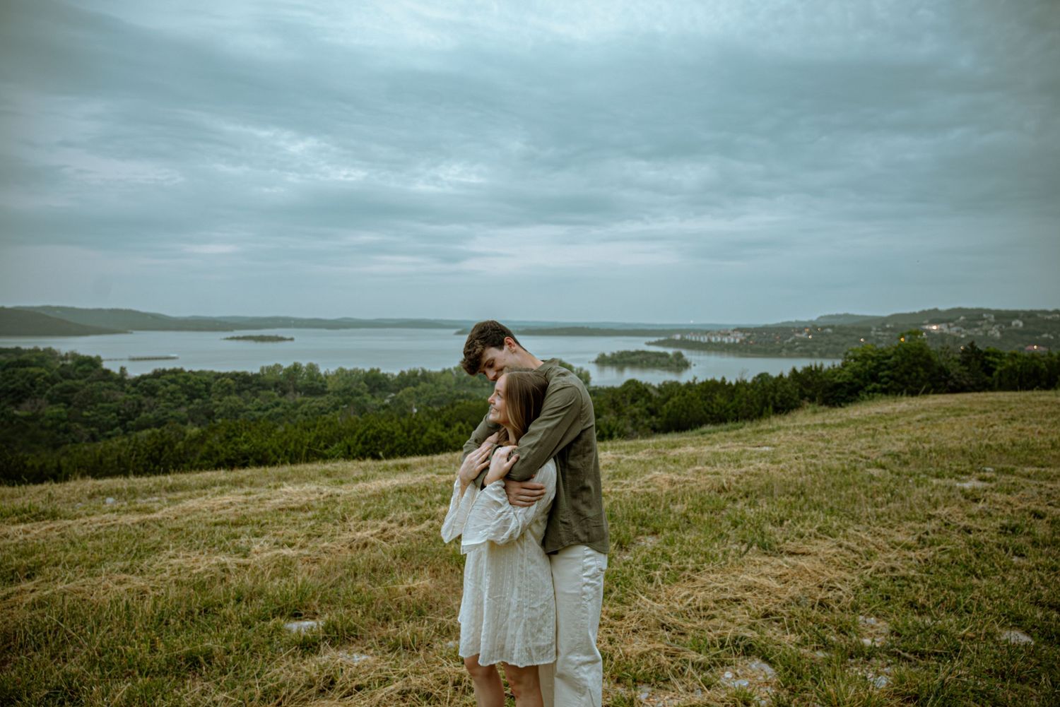 A couple embraces romantically on a grassy hillside overlooking a scenic lake under a cloudy sky.