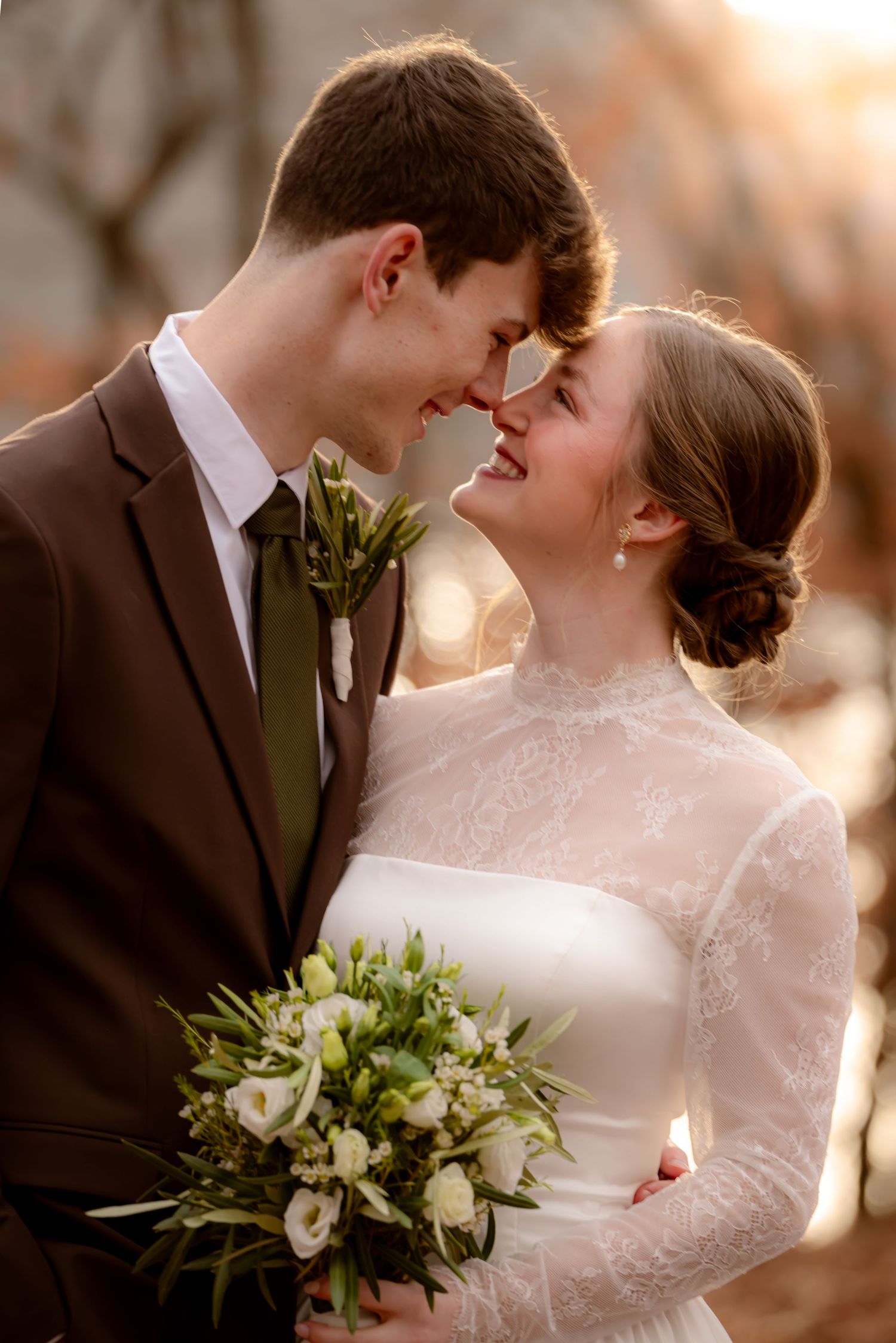 A bride and groom share a romantic moment, touching foreheads, as she holds a white floral bouquet in golden sunlight.