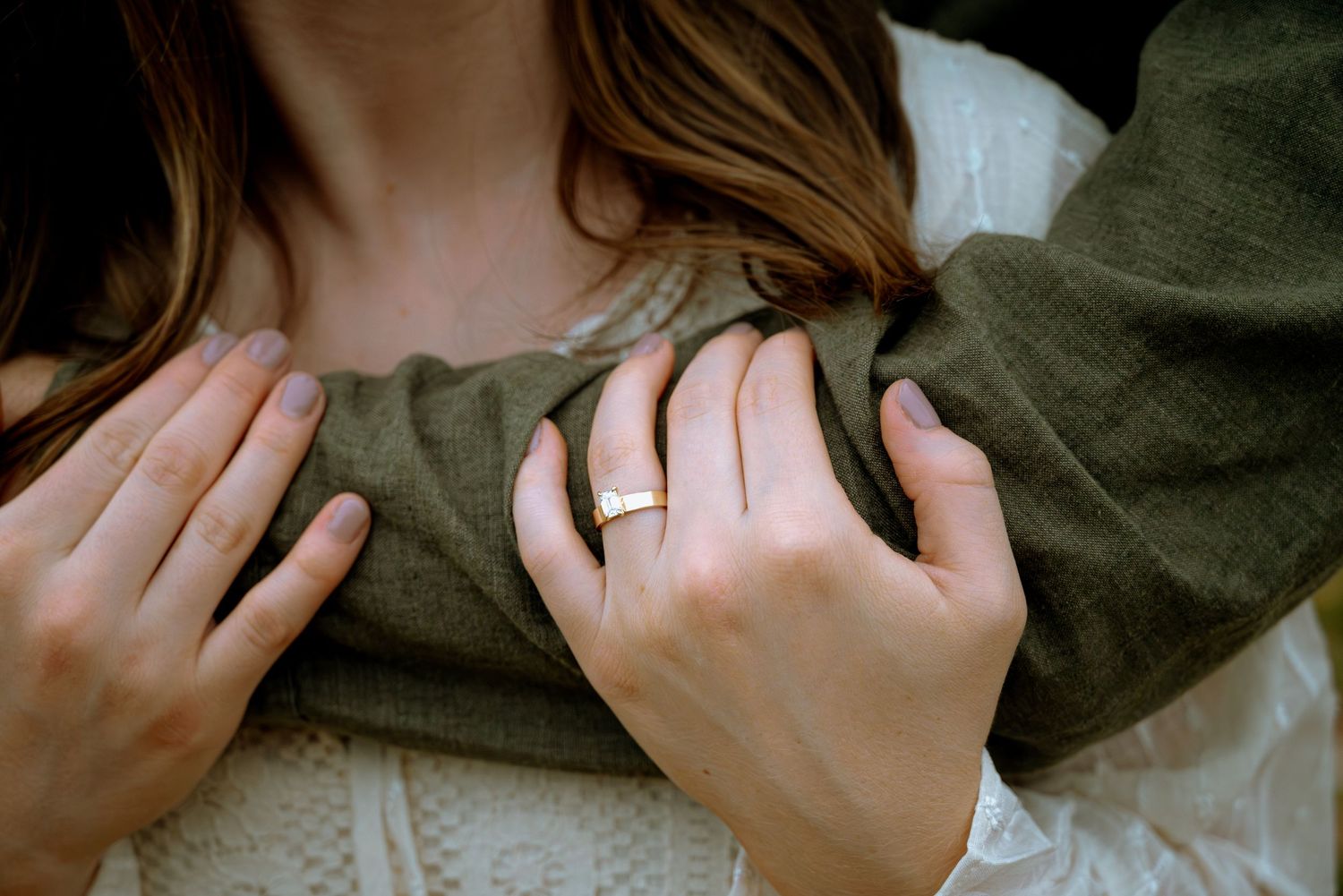 Close-up of a woman's hands wearing a gold engagement ring, clutching a man's olive green jacket.