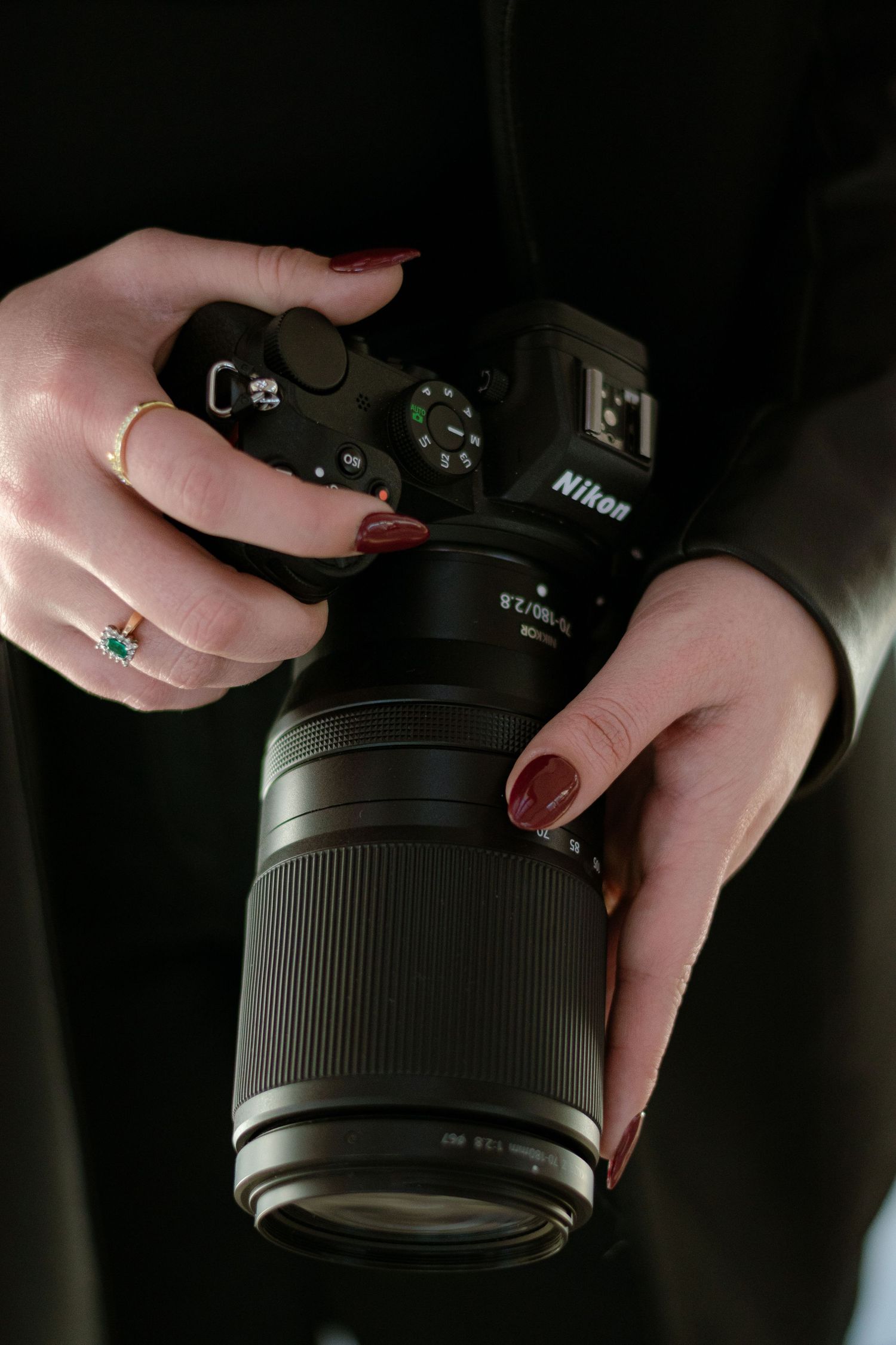 Close-up of woman's hands with red nails and gold rings holding a Nikon DSLR camera with lens.