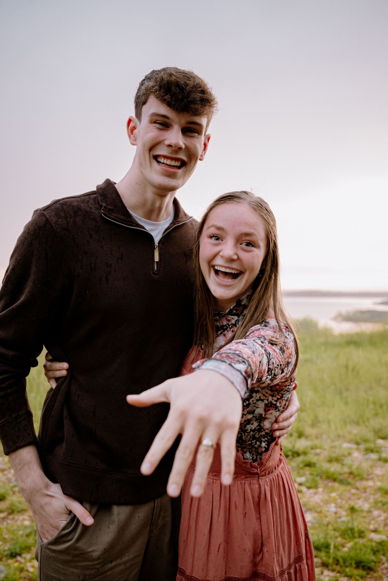 Excited newly engaged couple showing off engagement ring while smiling outdoors near a lake at dusk.
