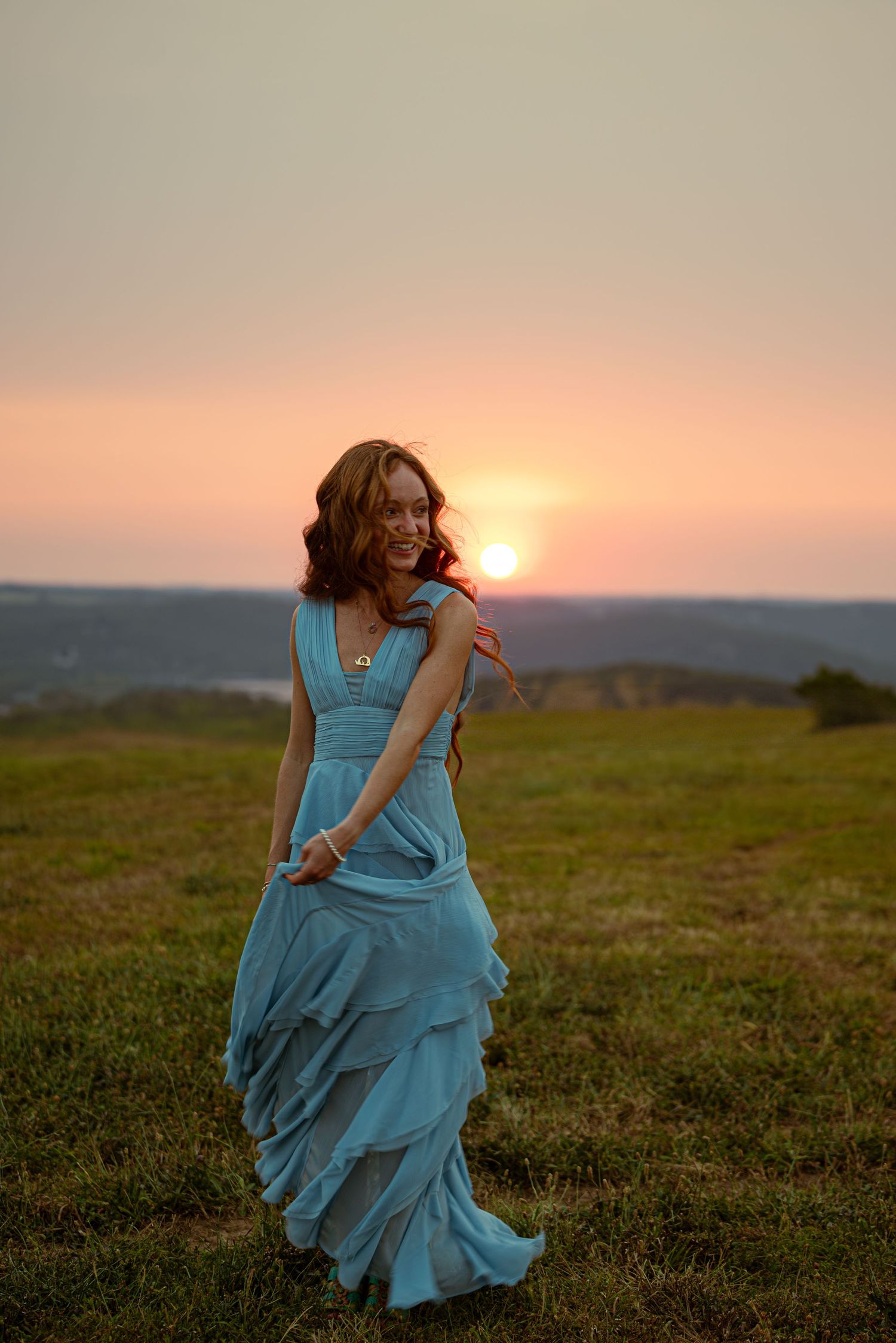Smiling woman in elegant blue ruffled gown twirling in grassy field at golden sunset.