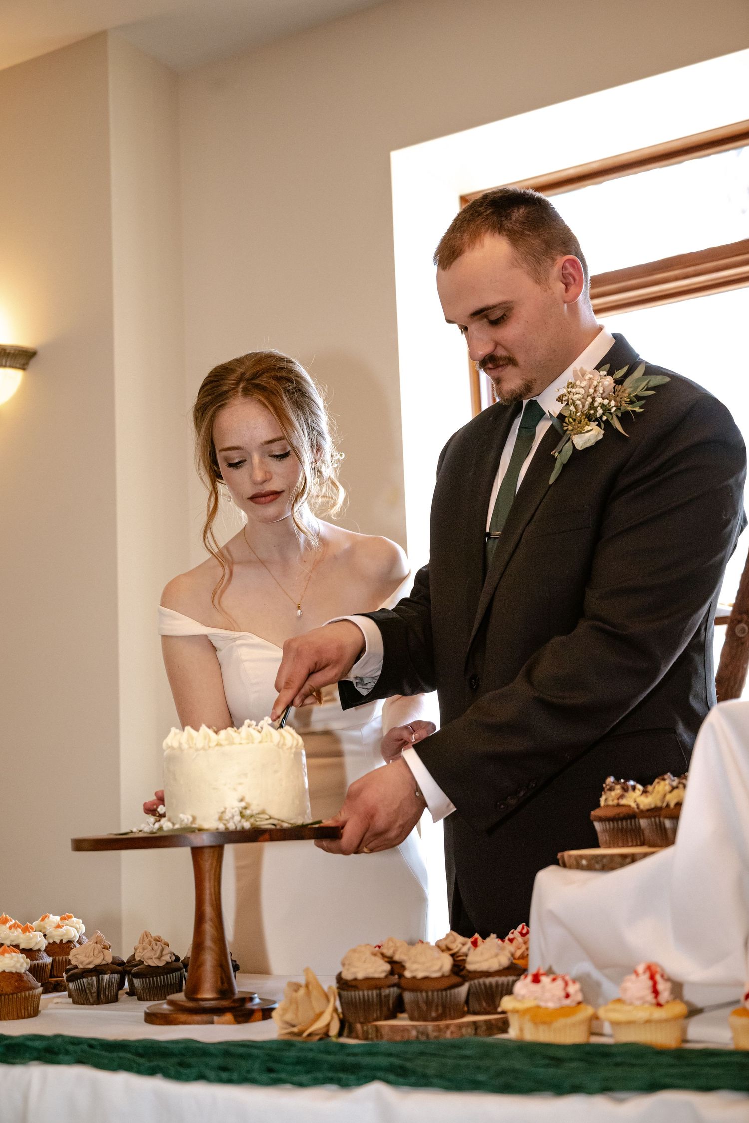Bride and groom cutting their white wedding cake at the chapel food bar, surrounded by wedding cookies.