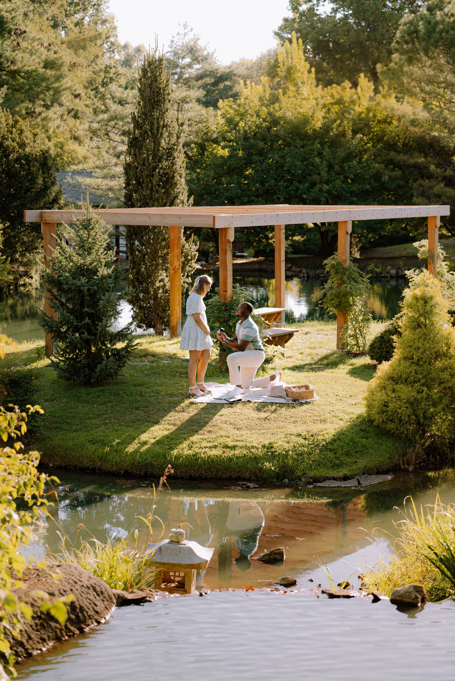 Interracial couple sharing a romantic proposal moment by a serene garden pond in golden sunlight.