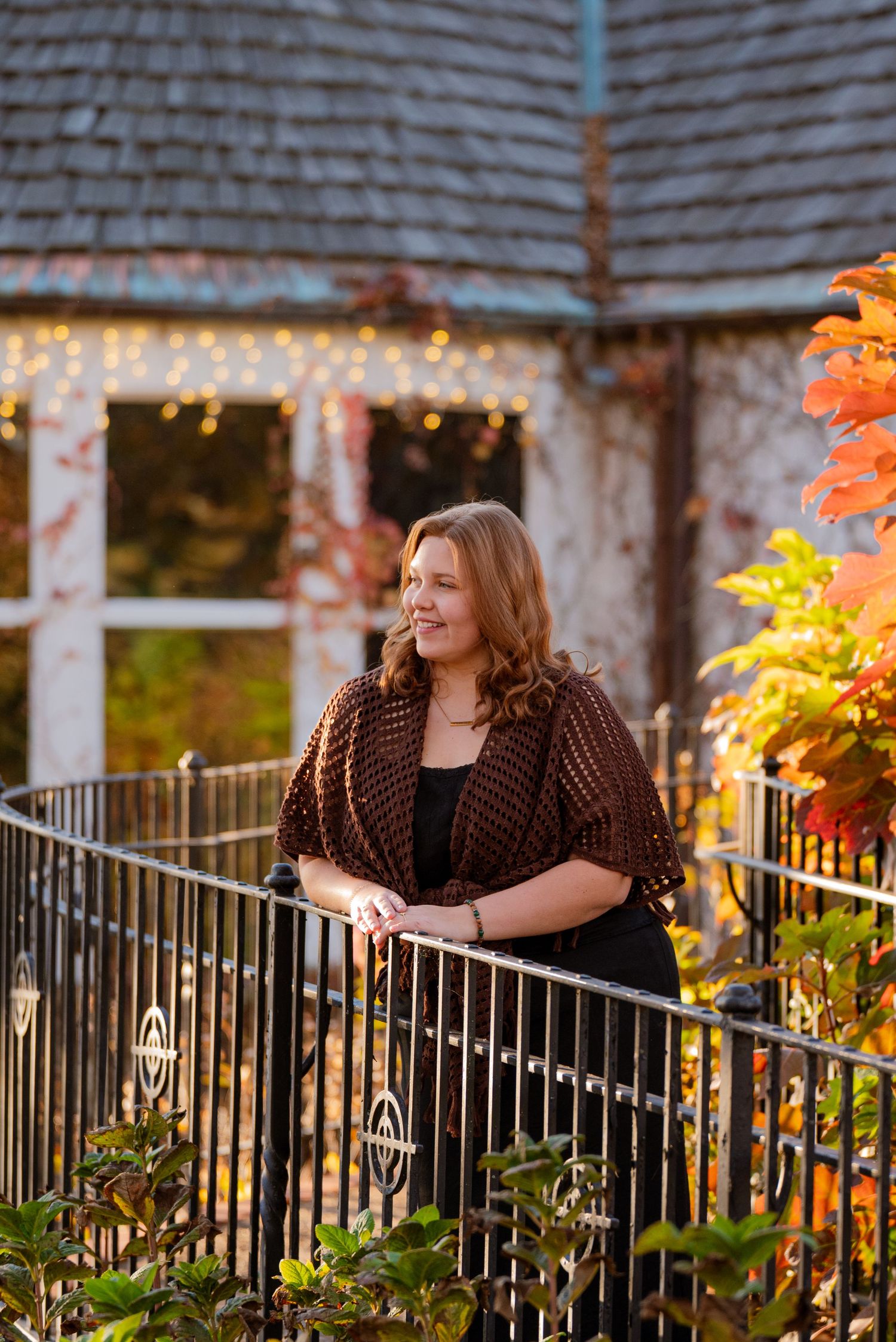Woman in brown crochet shawl leaning on iron railing surrounded by autumn foliage and fairy lights.