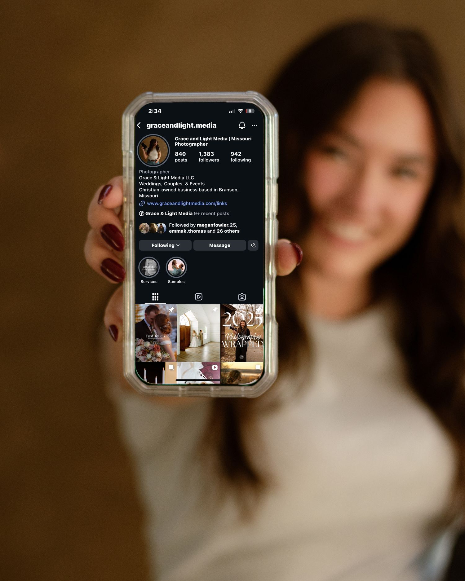 Smiling woman holding smartphone displaying Grace and Light Media photography Instagram profile page.