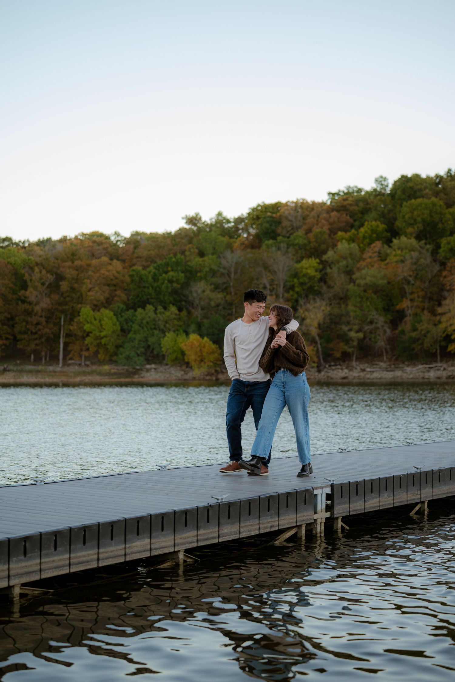 Couple sharing a romantic moment on a wooden dock by a calm lake surrounded by autumn trees.