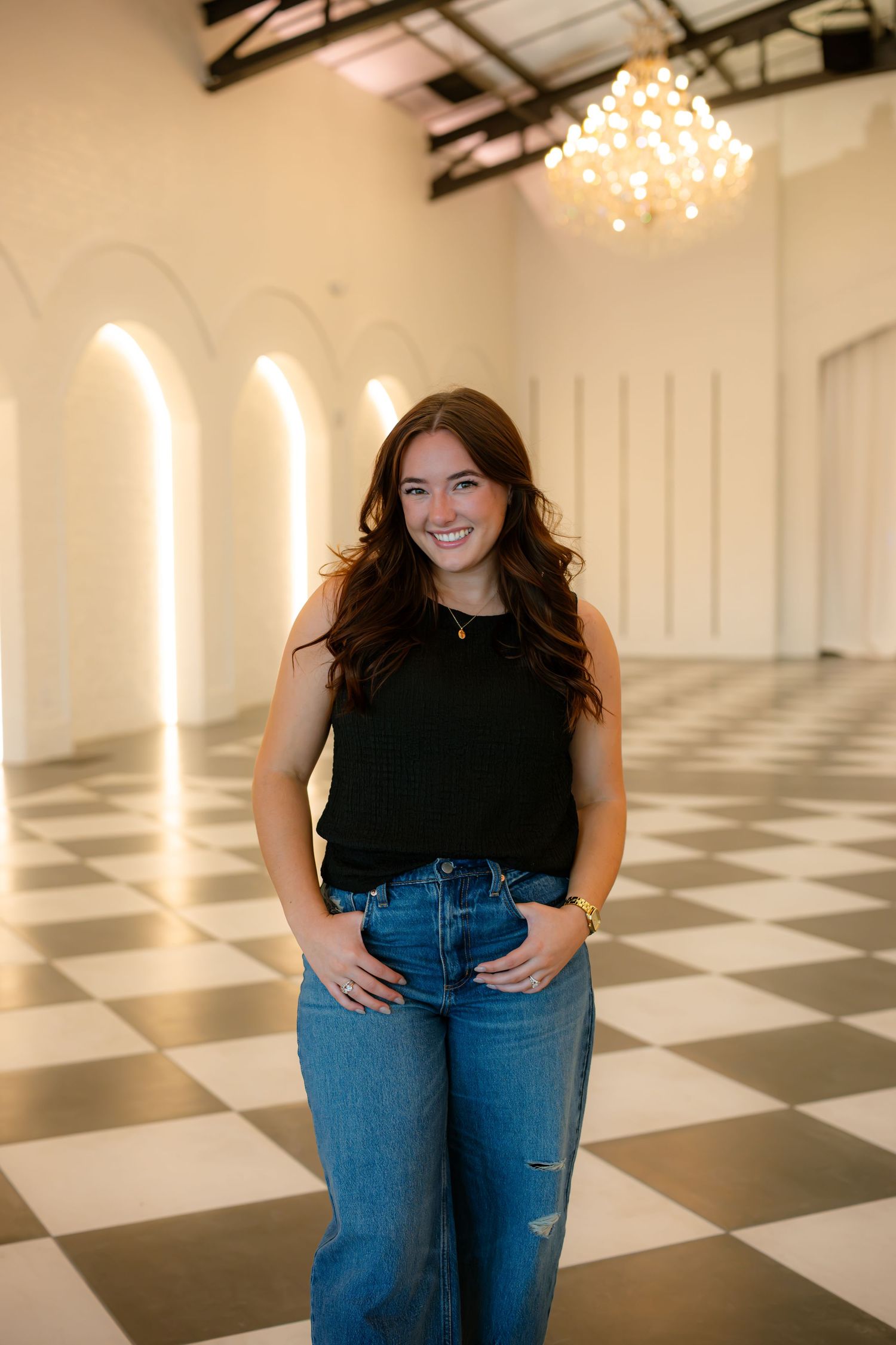 Young woman in black sleeveless top and ripped jeans smiling in elegant white venue with checkered floor.