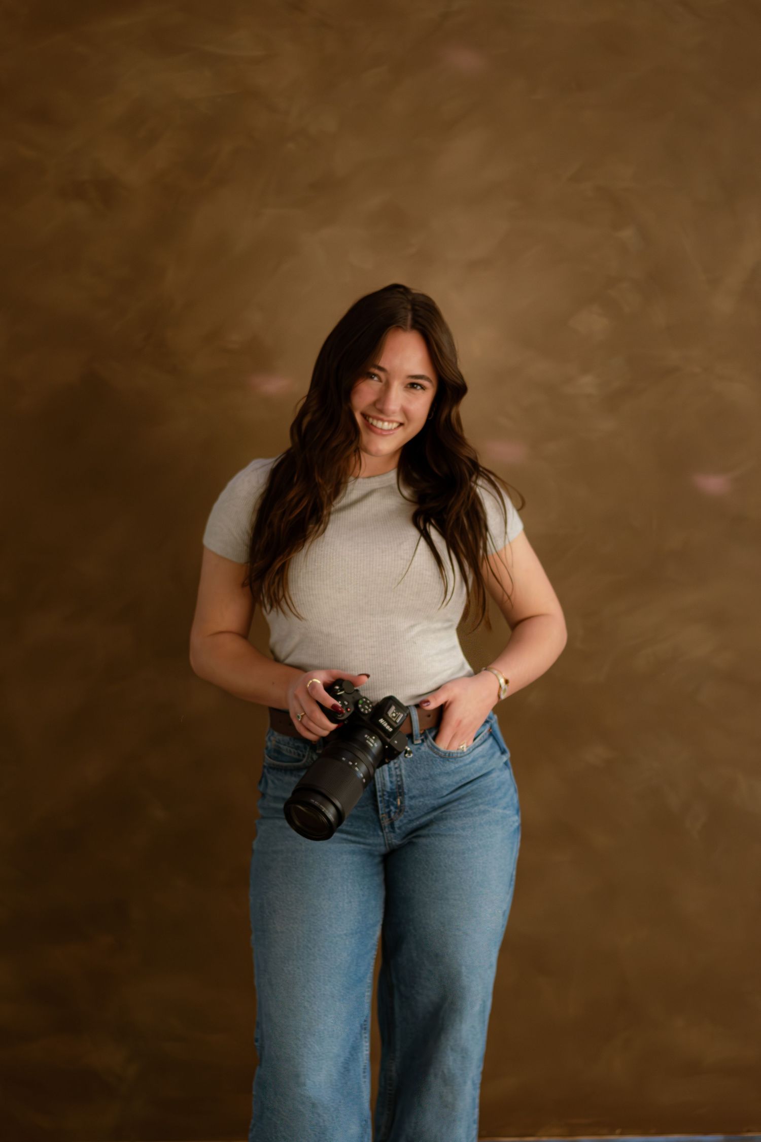 Smiling female photographer holding professional camera with hand in pocket against warm brown studio background.