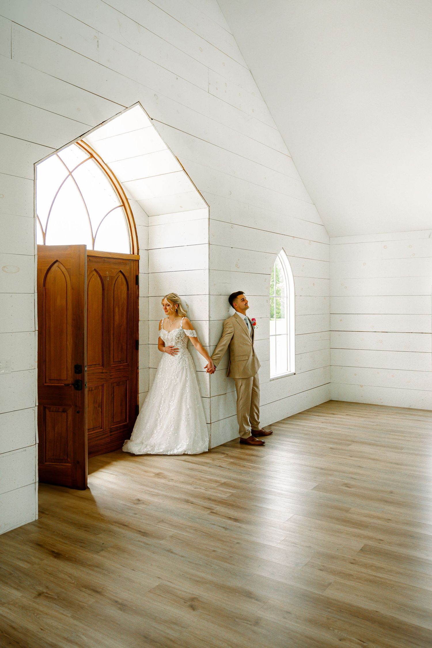 Bride and groom holding hands back-to-back inside a white chapel with arched wooden doors.