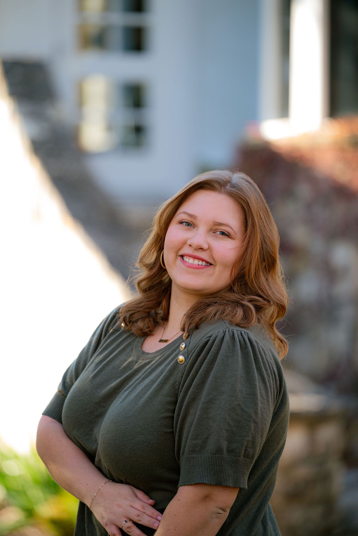 Smiling woman with auburn hair in olive green top posing outdoors in warm natural sunlight.