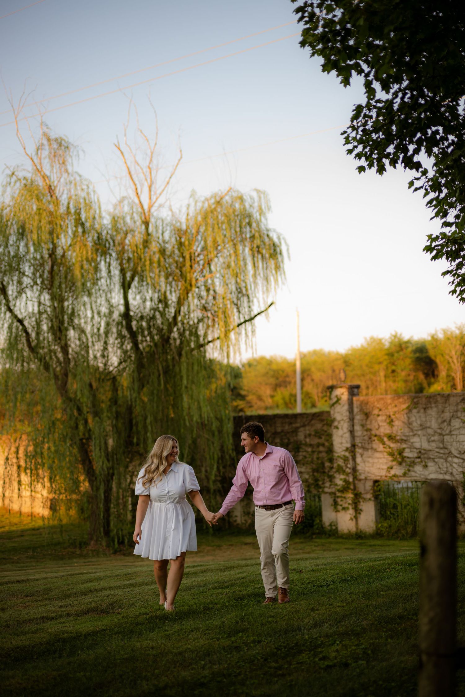 Couple holding hands walking on green grass near a willow tree during golden hour at an outdoor engagement session.