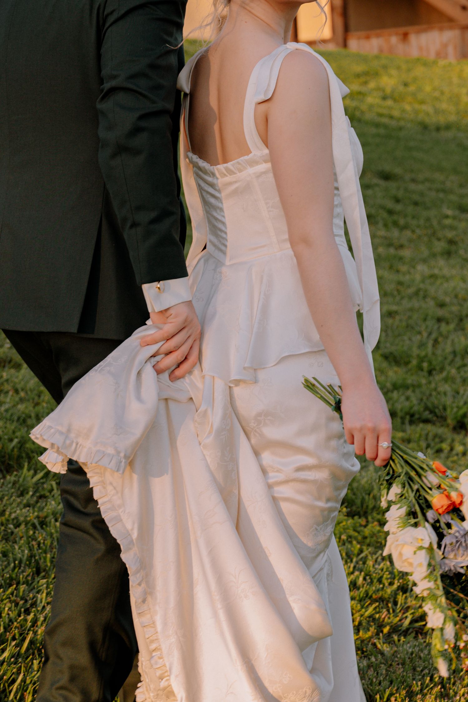 Bride in white corset wedding gown with groom in dark suit standing on green grass holding floral bouquet.