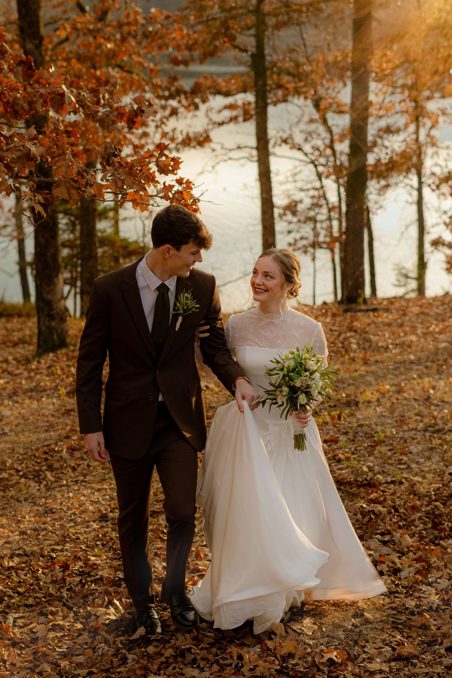 Bride and groom walk hand in hand through autumn forest with golden leaves and lake backdrop.