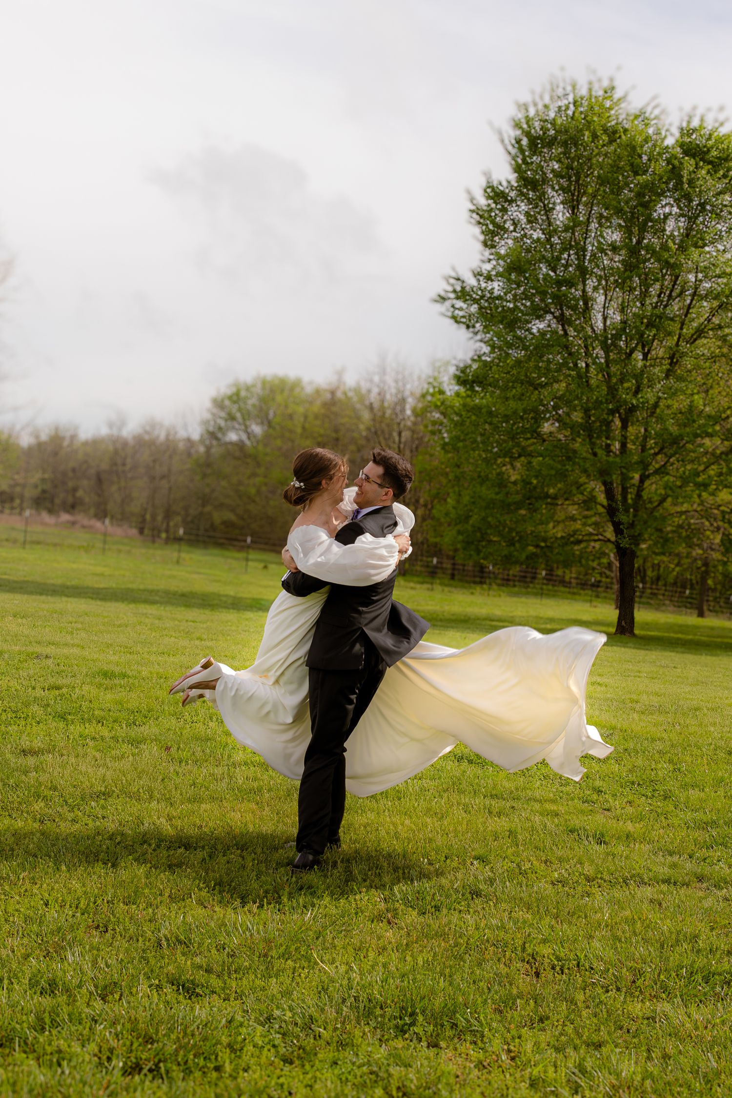 Groom lifts bride in white flowing dress outdoors on lush green lawn with trees in background on wedding day.