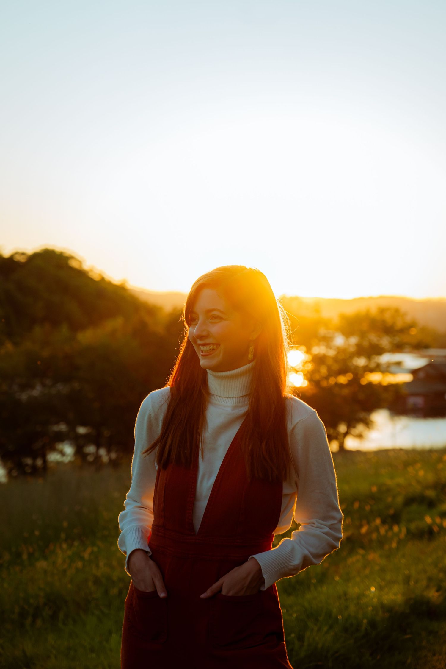 A smiling young woman in a white turtleneck and red vest stands outdoors during a golden sunset.