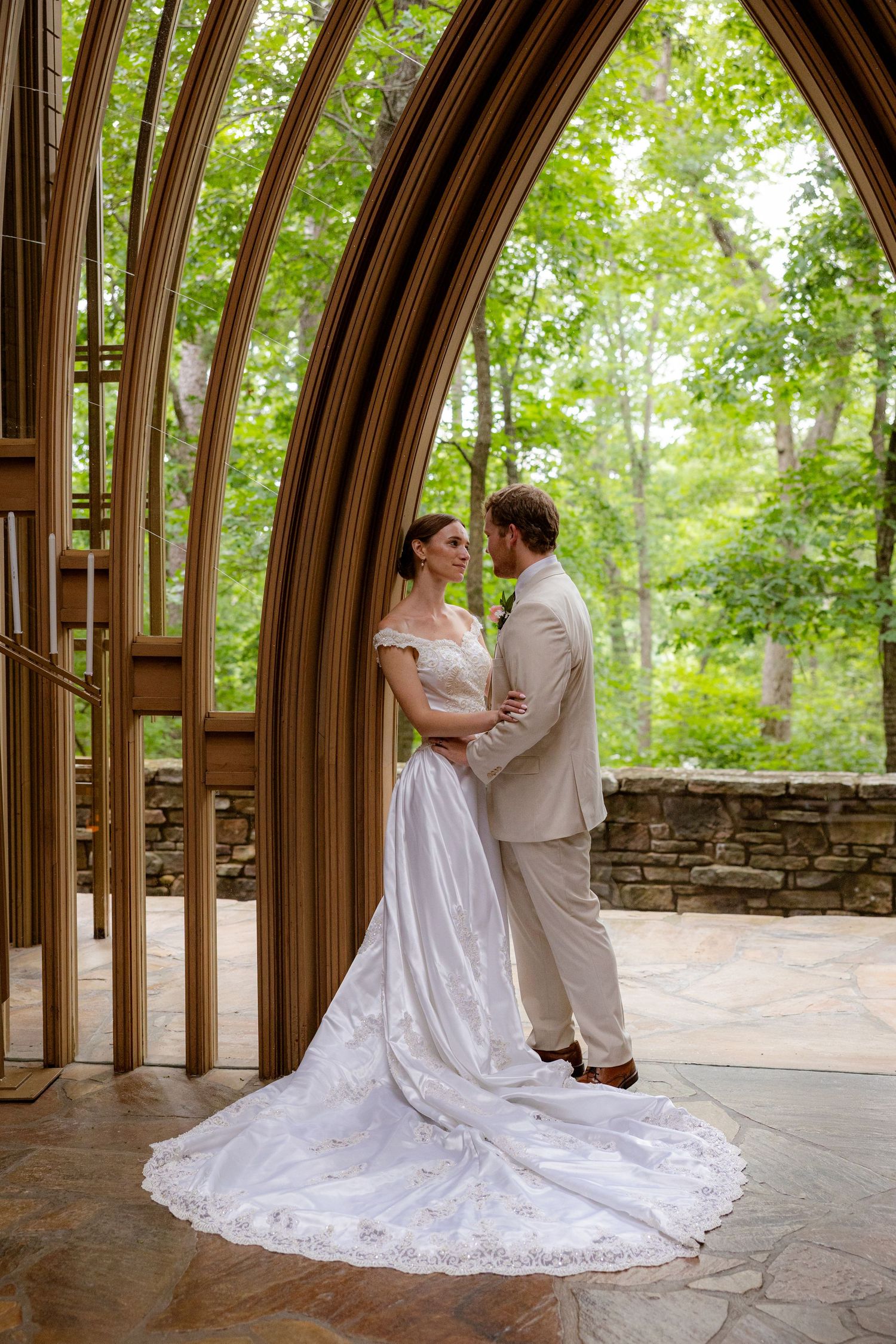 Bride and groom embracing under elegant wooden arch The Mildred B. Memorial chapel surrounded by lush green trees.