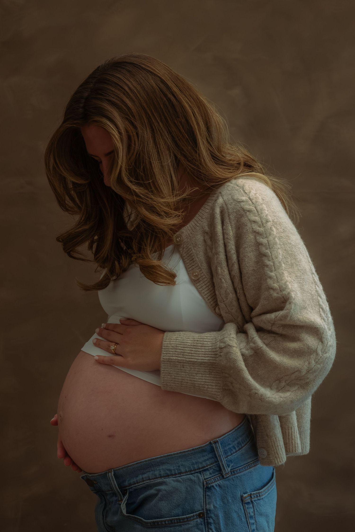 Pregnant woman in beige cardigan and jeans cradling her baby bump against a dark studio background.