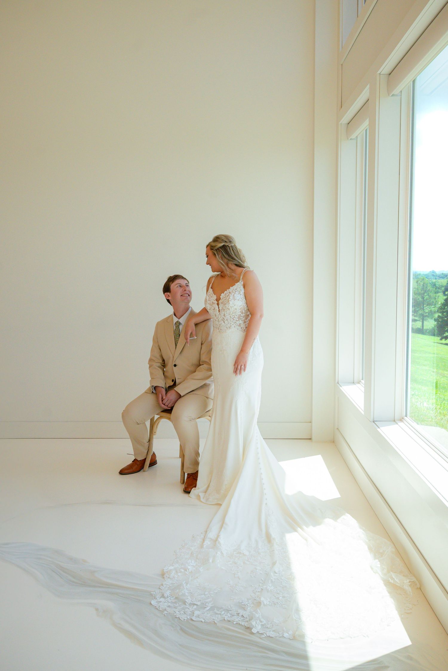 Bride in lace gown and groom in tan suit share a joyful moment by large windows in a bright white venue.