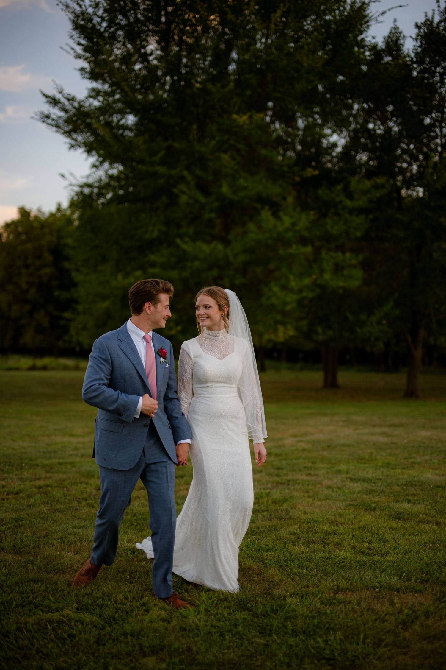 Bride and groom walk hand in hand across green lawn at sunset, she in white gown with veil, he in blue suit.