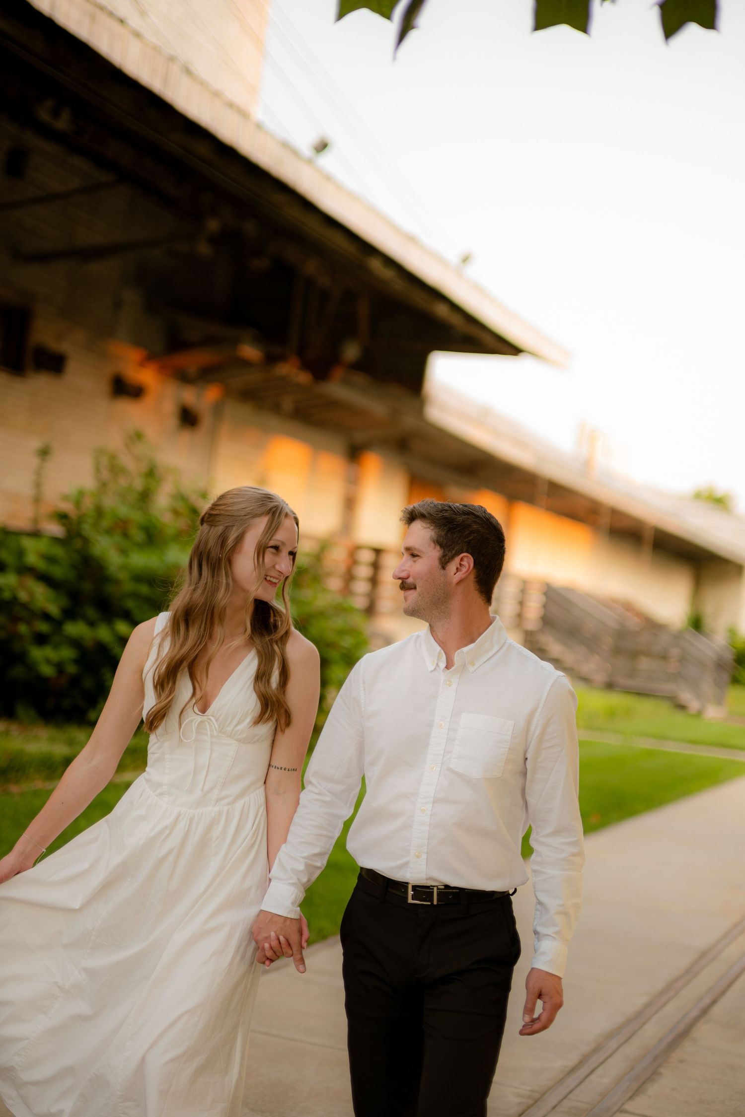 Couple in white attire walks hand-in-hand, smiling at each other outdoors near a rustic building at golden hour.