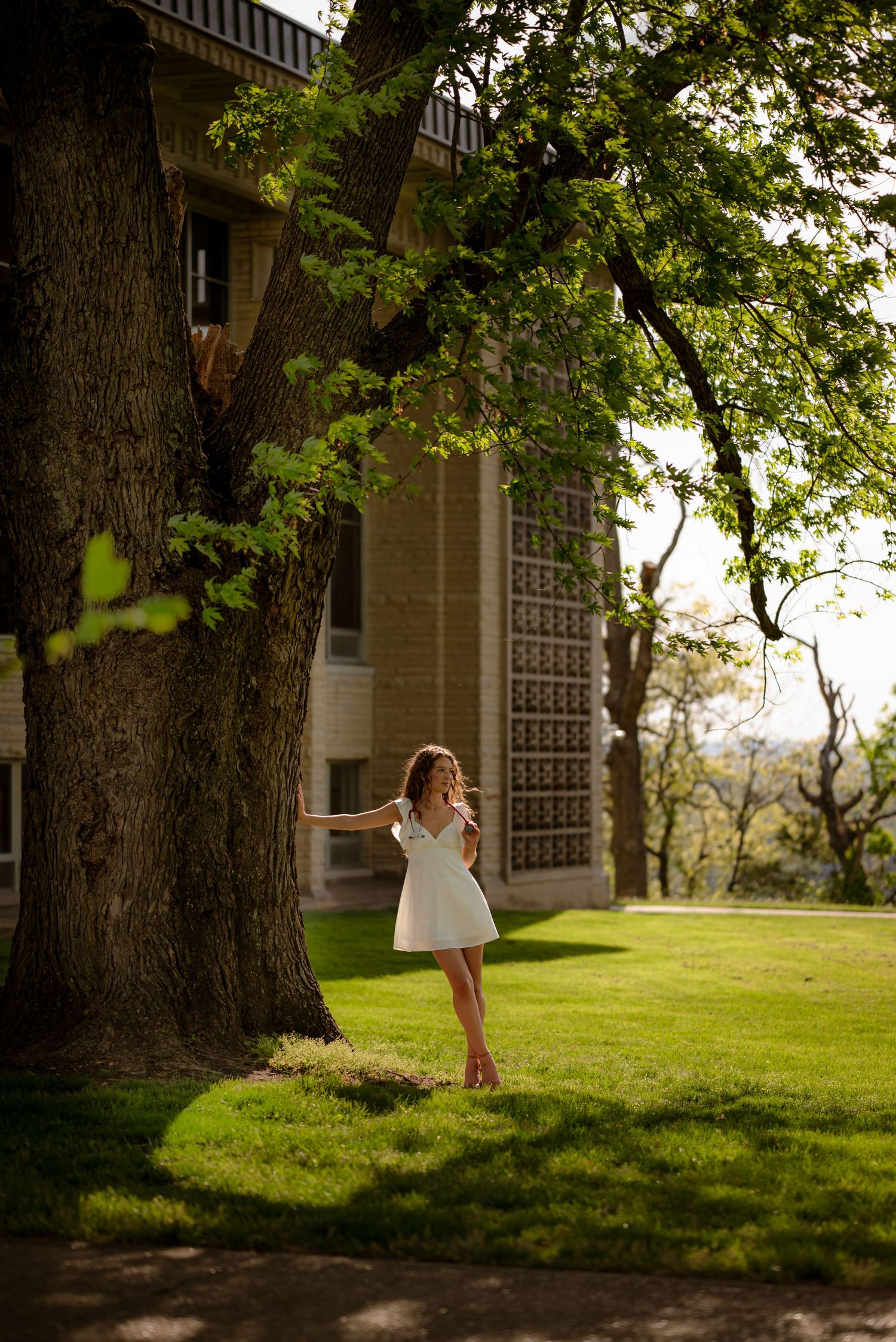 Young woman in white dress dancing near large tree on green lawn beside historic building on sunny day.