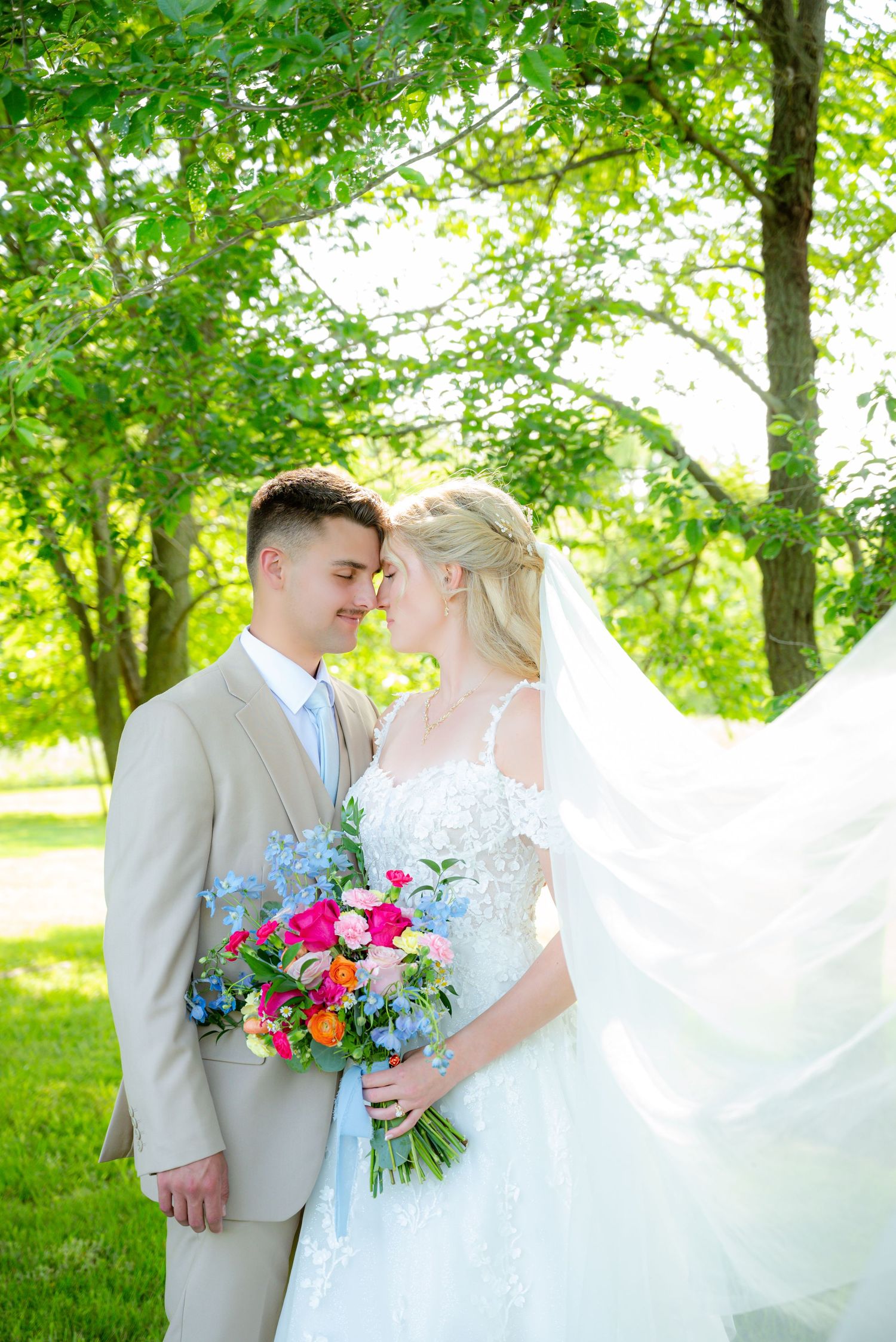 Bride and groom share a romantic nose-to-nose moment outdoors, her veil flowing, holding a vibrant pink and blue bouquet.