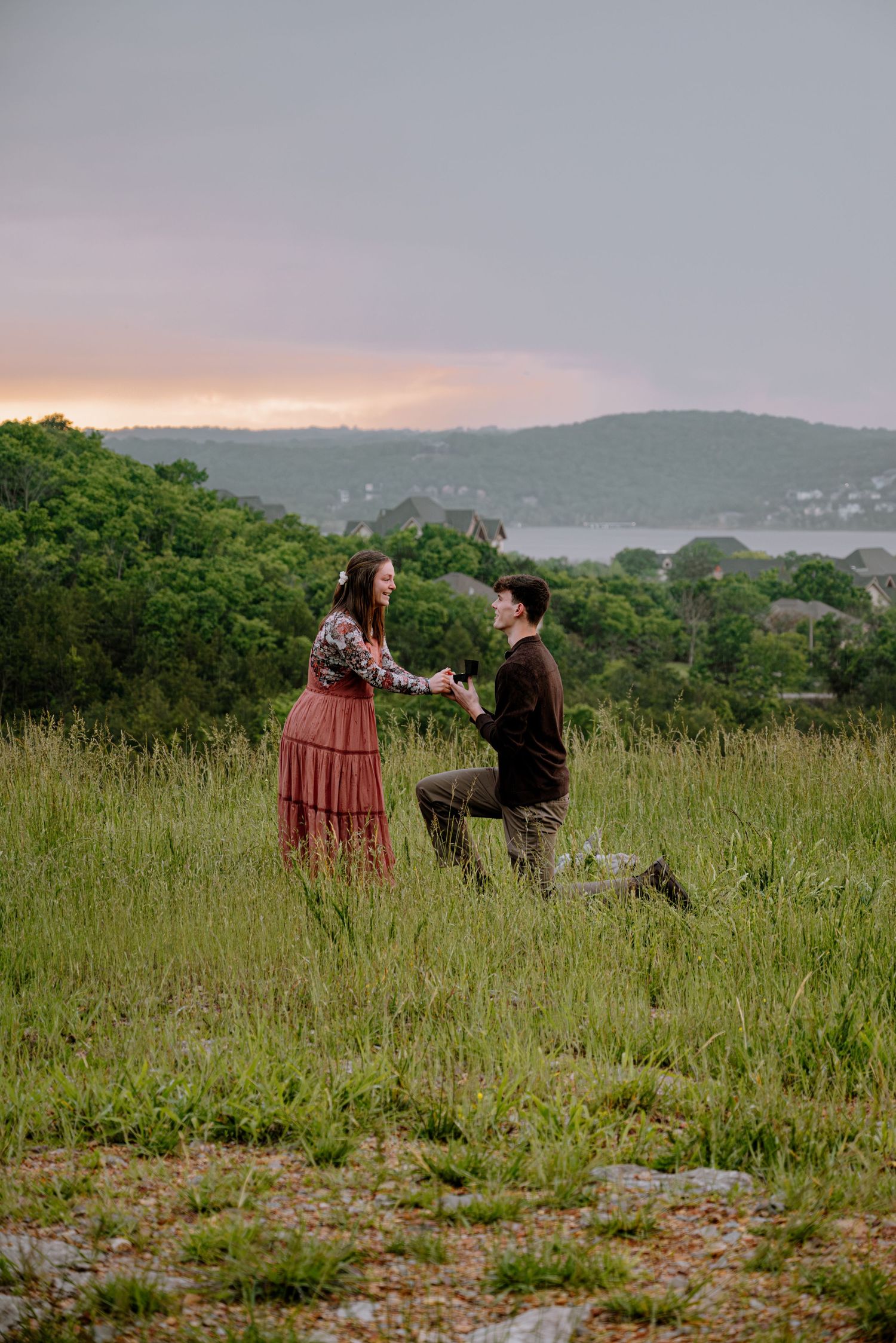 Man proposing on one knee to woman in red dress in a green field at sunset overlooking a lake.