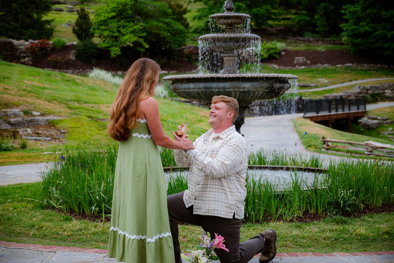 Man proposes on one knee to woman in green dress near stone fountain in lush garden setting.
