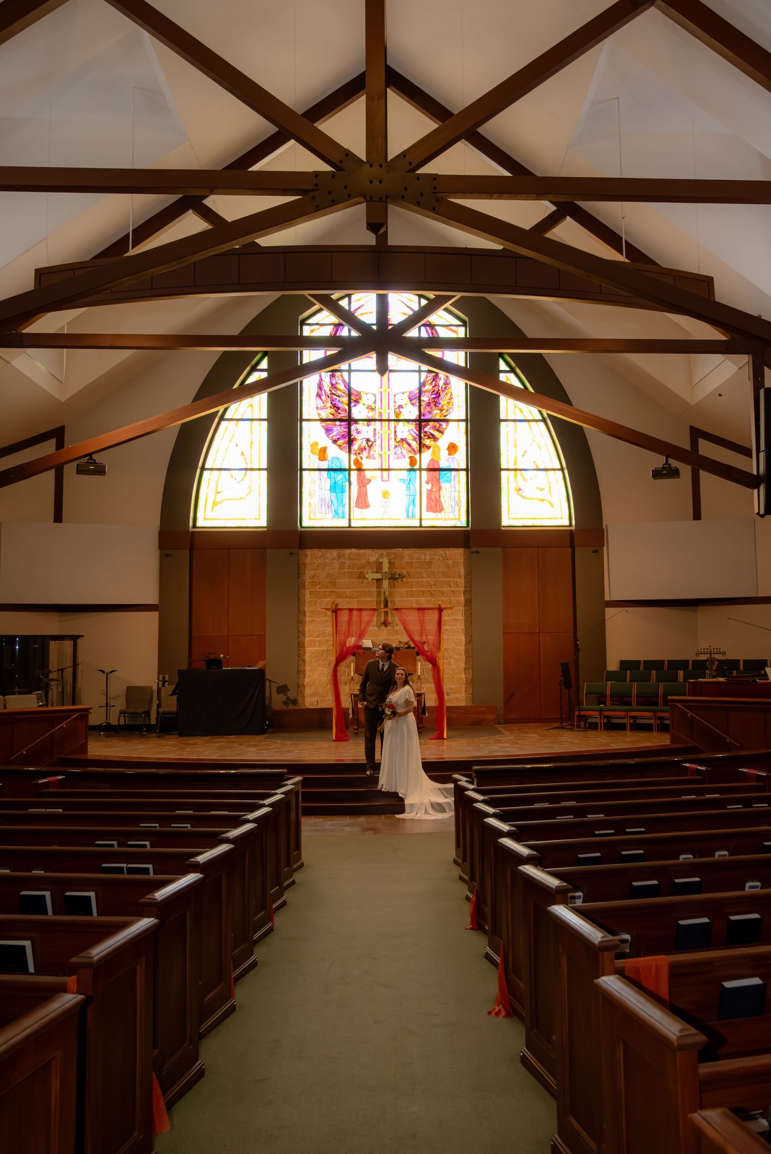 Bride and groom stand at the altar of a church with stunning stained glass windows and wooden pews.