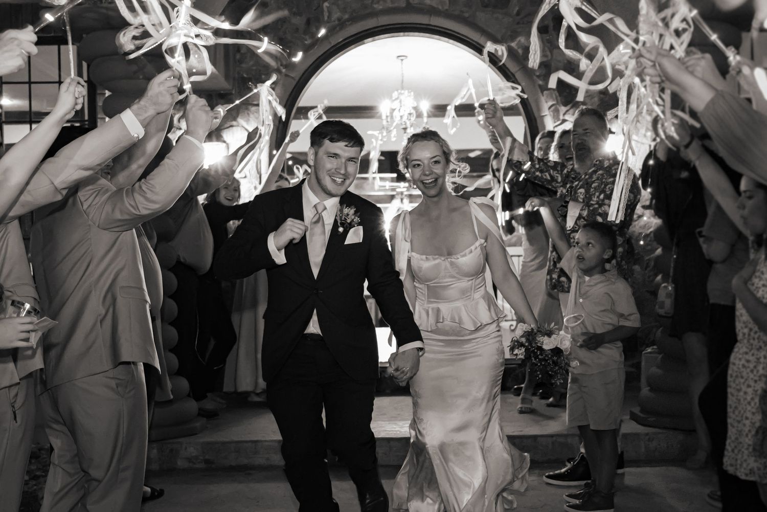 Bride and groom exiting their wedding reception out the front door of their wedding venue, surrounded by ribbons & friends.