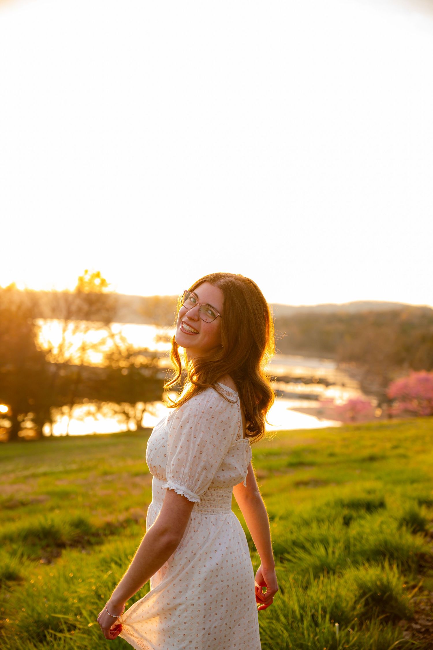 Woman in white dress laughs joyfully at golden hour sunset near a scenic lakeside hillside.