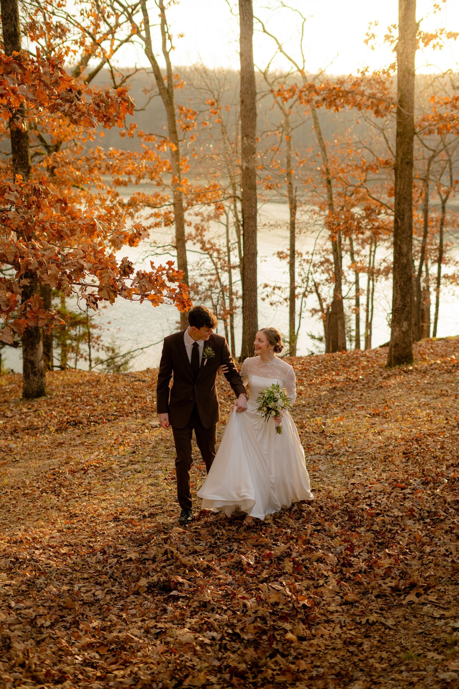Bride and groom walk hand-in-hand through autumn forest with golden leaves and lake backdrop at sunset.